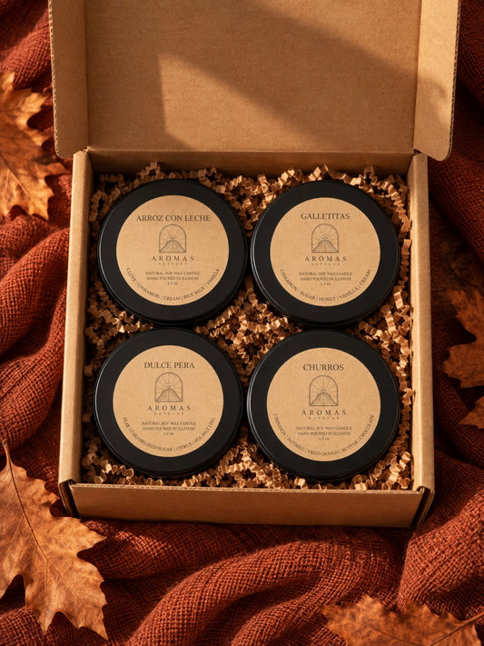 Open kraft gift box containing four Aromas Aztecas 1.5 oz fall discovery candles: Arroz con Leche, Galletitas, Dulce Pera, and Churros, nestled in kraft crinkle paper. The box is surrounded by terracotta fabric and autumn leaves.
