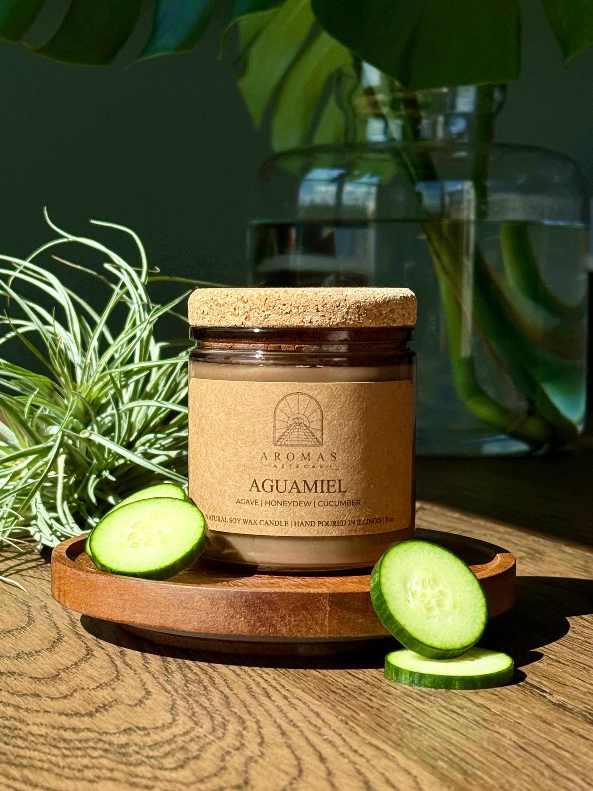 Aromas Aztecas Aguamiel soy candle in an amber glass vessel with a cork lid on a wooden textured surface with sliced cucumbers with leaves and a clear vase with water.