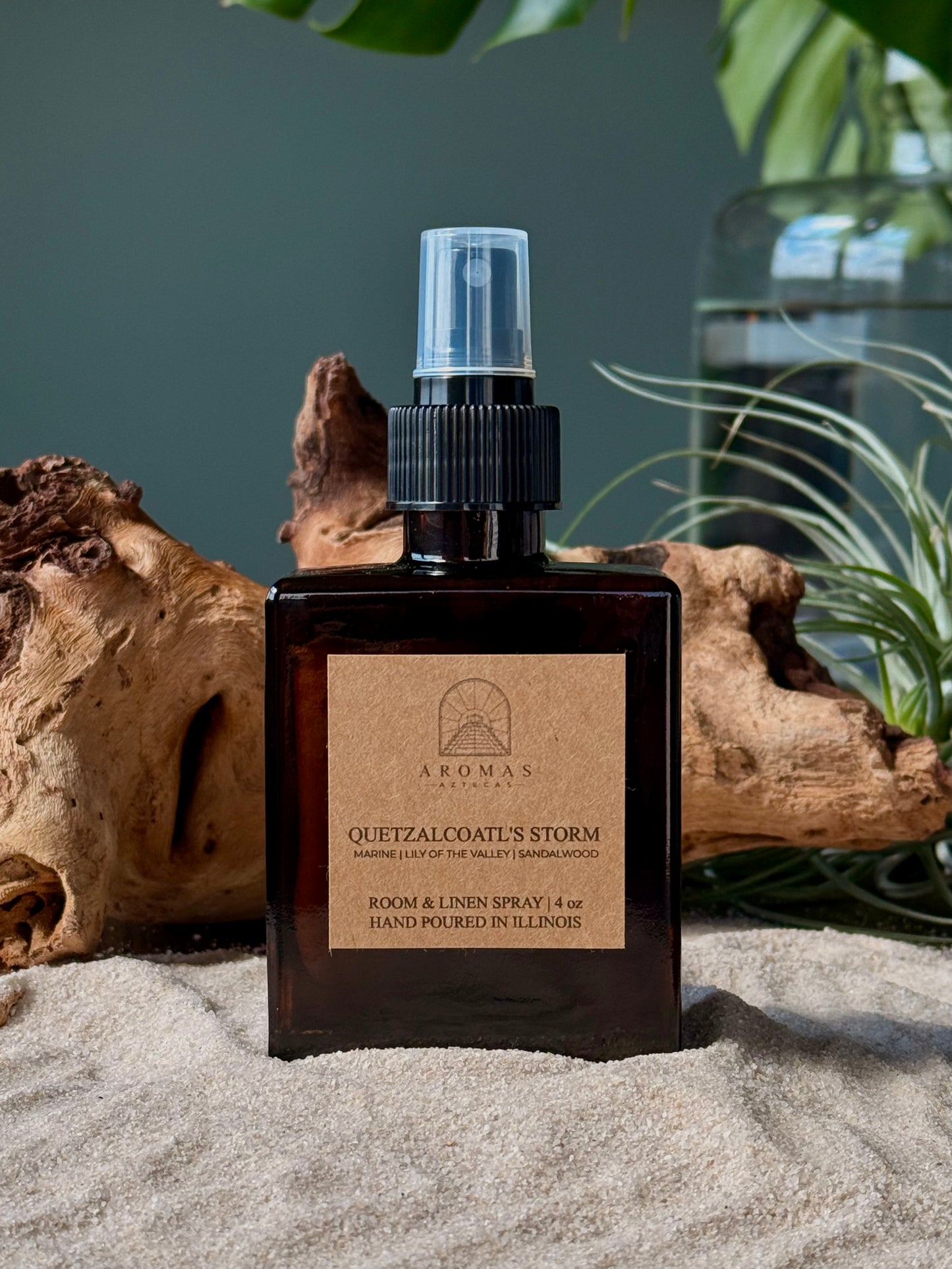 Aromas Aztecas Quetzalcoatl's Storm room and linen spray in an amber glass bottle on a sandy surface with wood and leaves.
