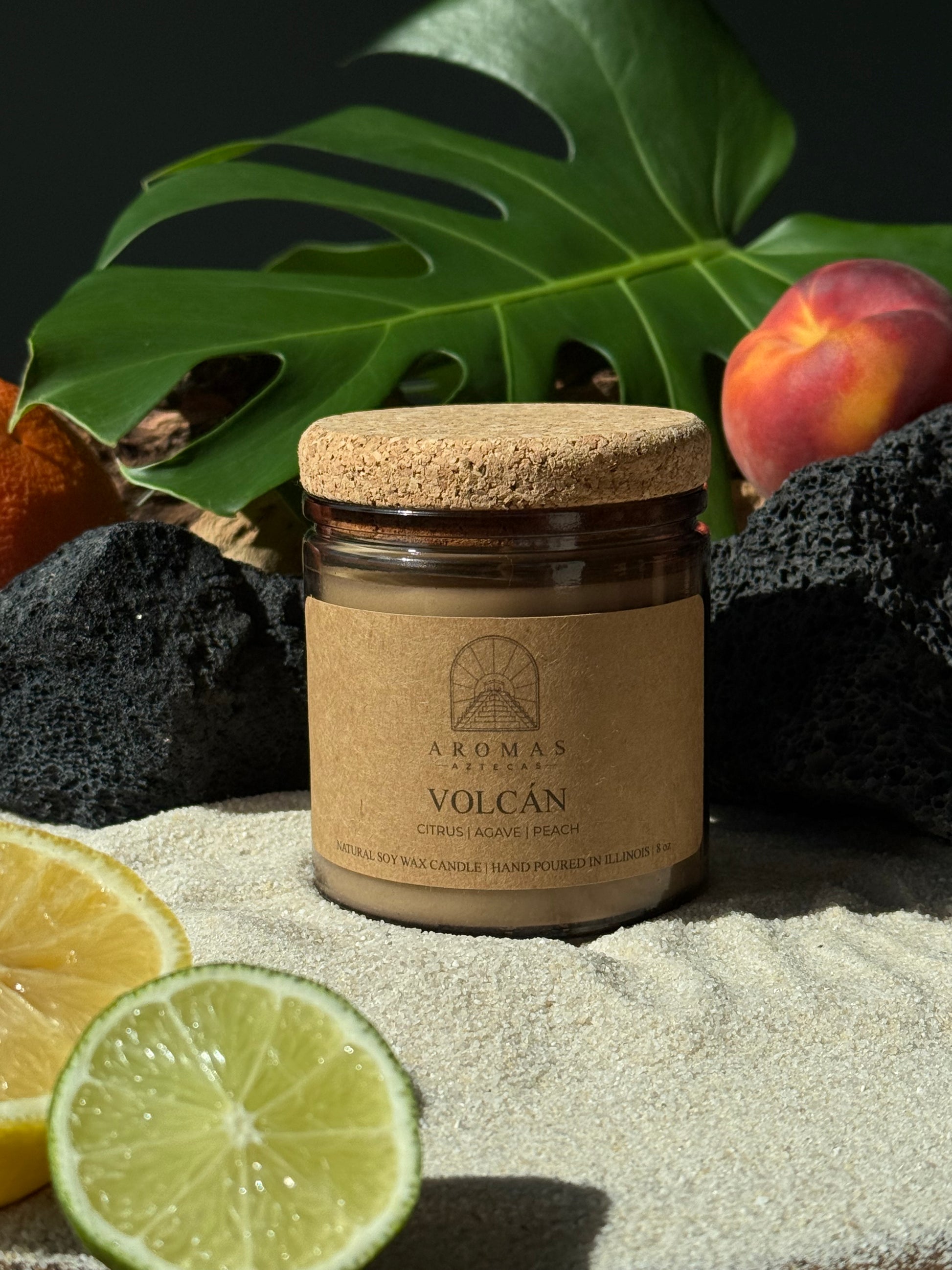 Aromas Aztecas Volcán soy candle in an amber glass vessel with a cork lid on a sandy surface surrounded by black lava rock, lime, lemon, peaches and leaves.