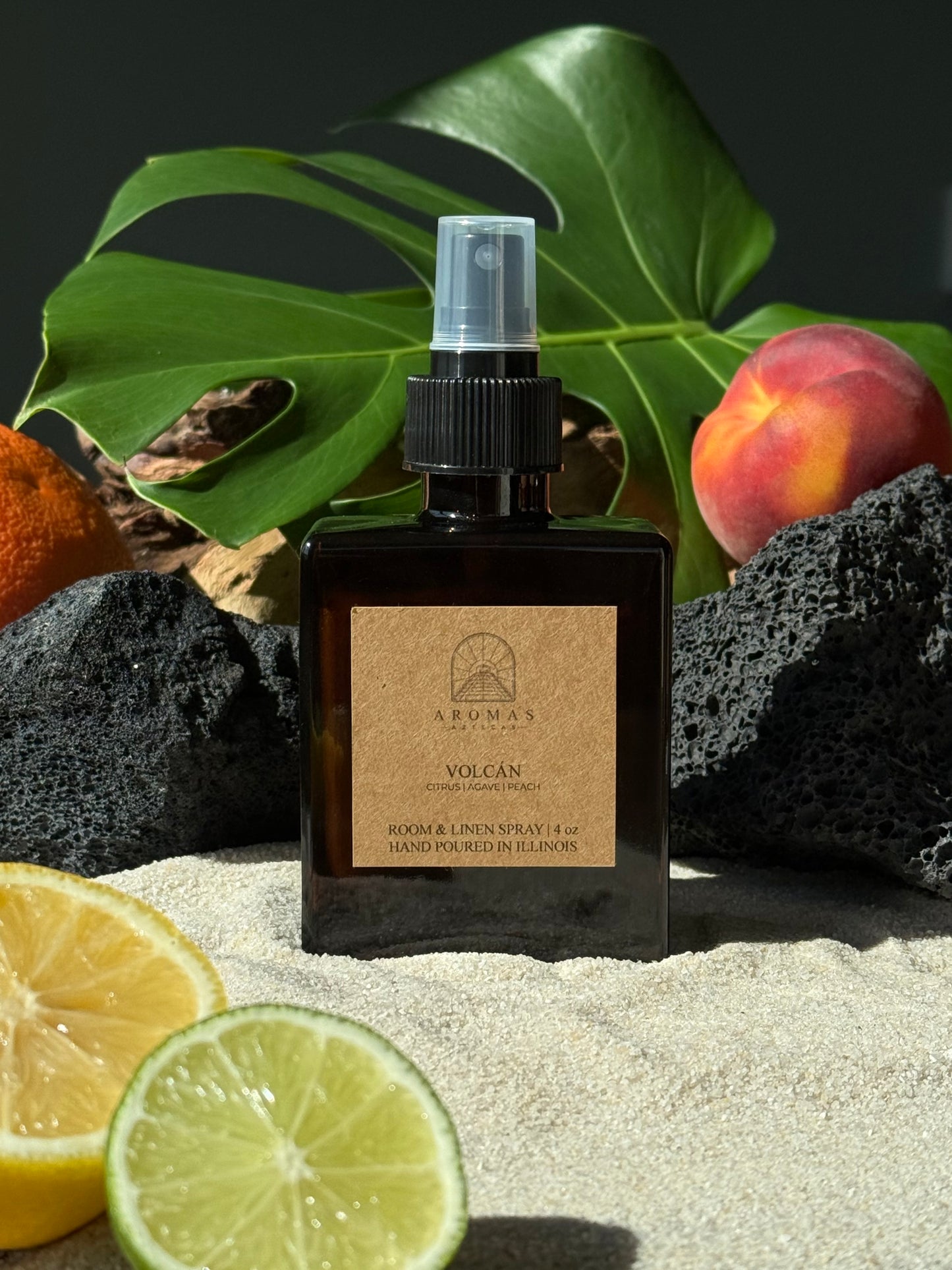 Aromas Aztecas Volcán room and linen spray in an amber glass bottle on a sandy surface with black lava rock, lime, lemon, peaches, and leaves.