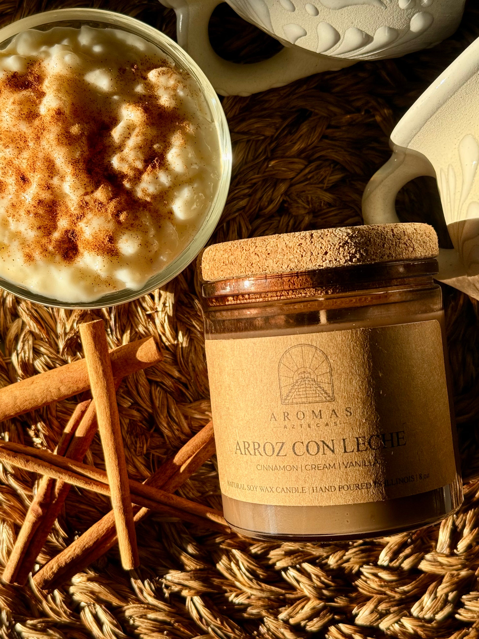 Aromas Aztecas Arroz con Leche soy candle in an amber glass vessel with a cork lid laying on a woven textured surface next to cinnamon sticks and a cup of rice pudding.