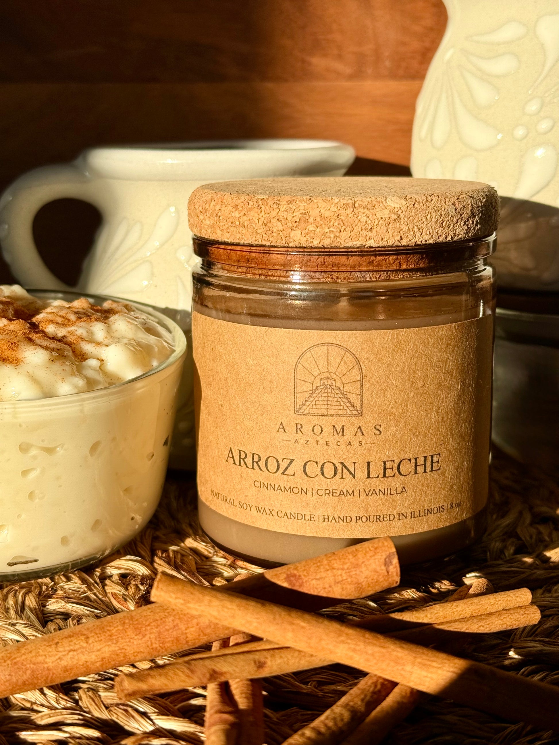 Aromas Aztecas Arroz con Leche soy candle in an amber glass vessel with a cork lid on a woven textured surface behind cinnamon sticks and next to a cup of rice pudding.