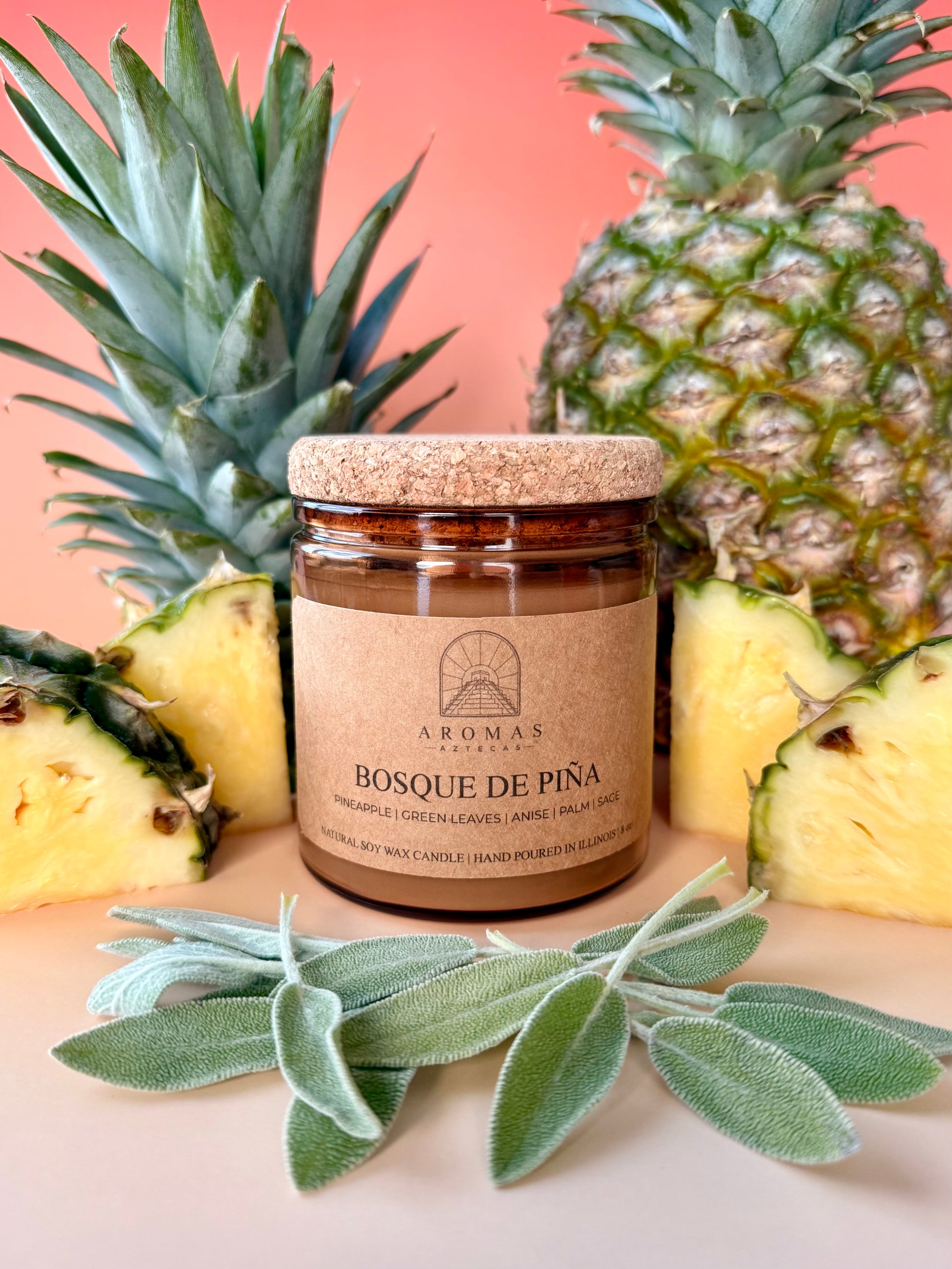 Aromas Aztecas Bosque de Piña soy candle in an amber glass vessel with a cork lid on a sunset pink background surrounded by pineapple and sage.