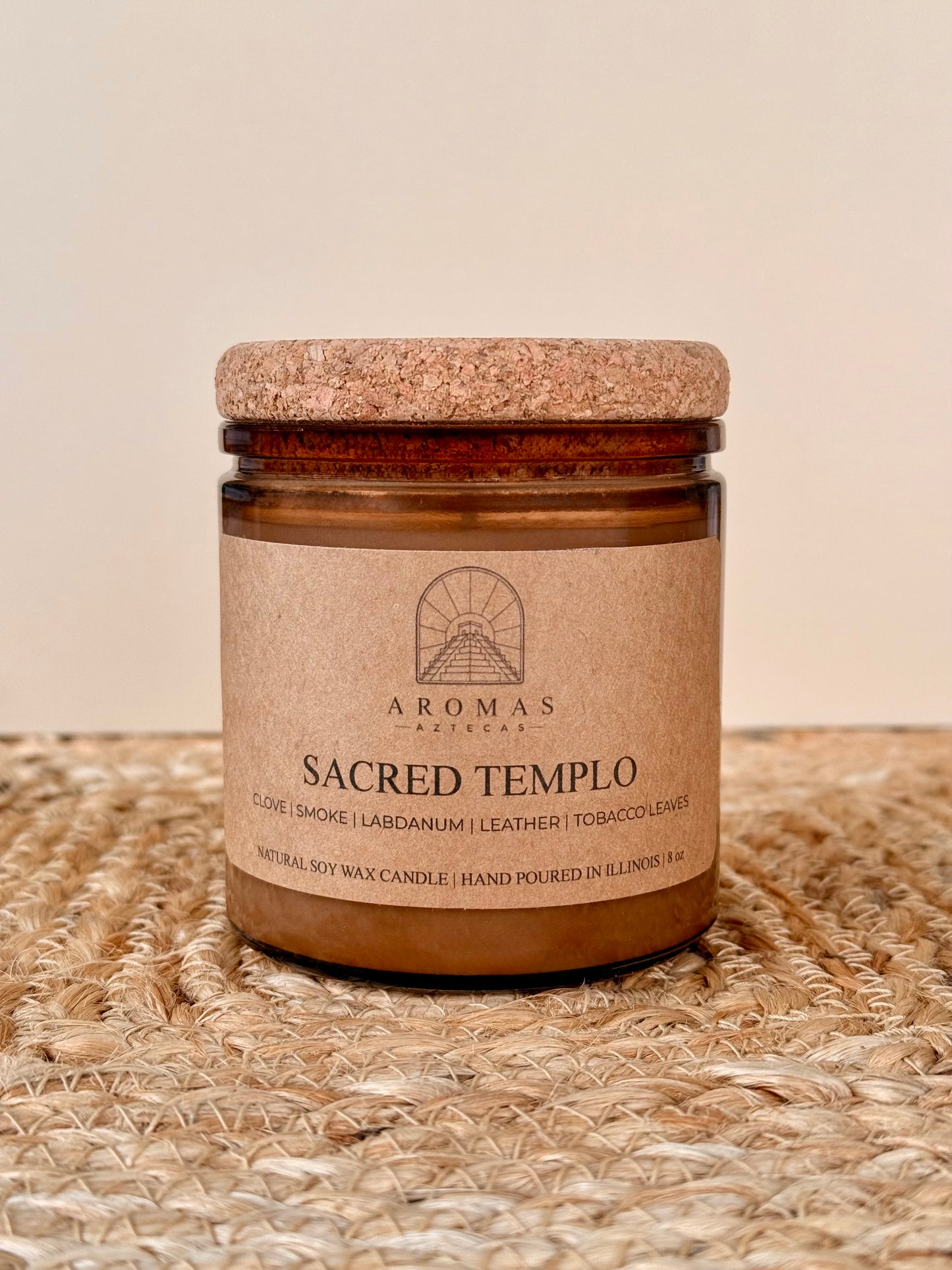 Aromas Aztecas Sacred Templo soy candle in an amber glass vessel with a cork lid on a woven textured surface.