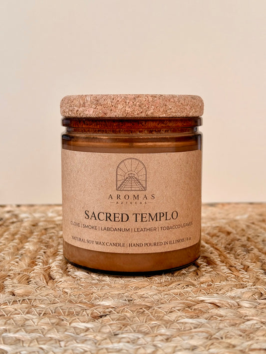 Aromas Aztecas Sacred Templo soy candle in an amber glass vessel with a cork lid on a woven textured surface.