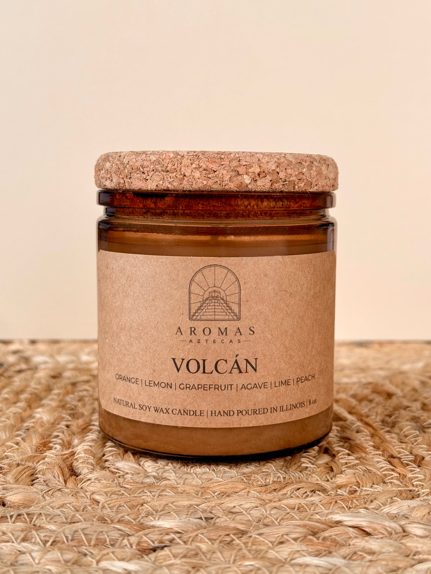 Aromas Aztecas Volcán soy candle in an amber glass vessel with a cork lid on a woven textured surface.