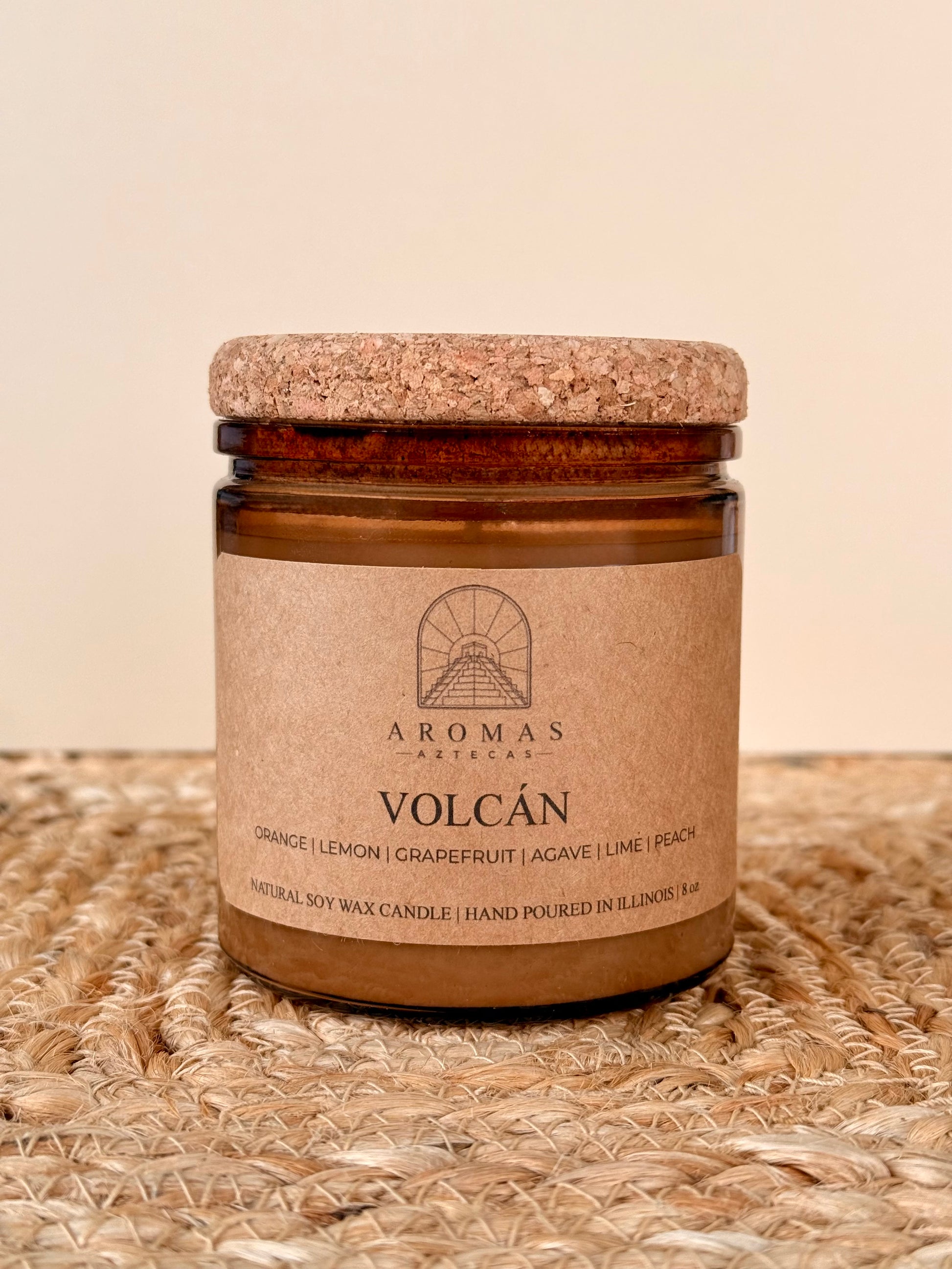 Aromas Aztecas Volcán soy candle in an amber glass vessel with a cork lid on a woven textured surface.