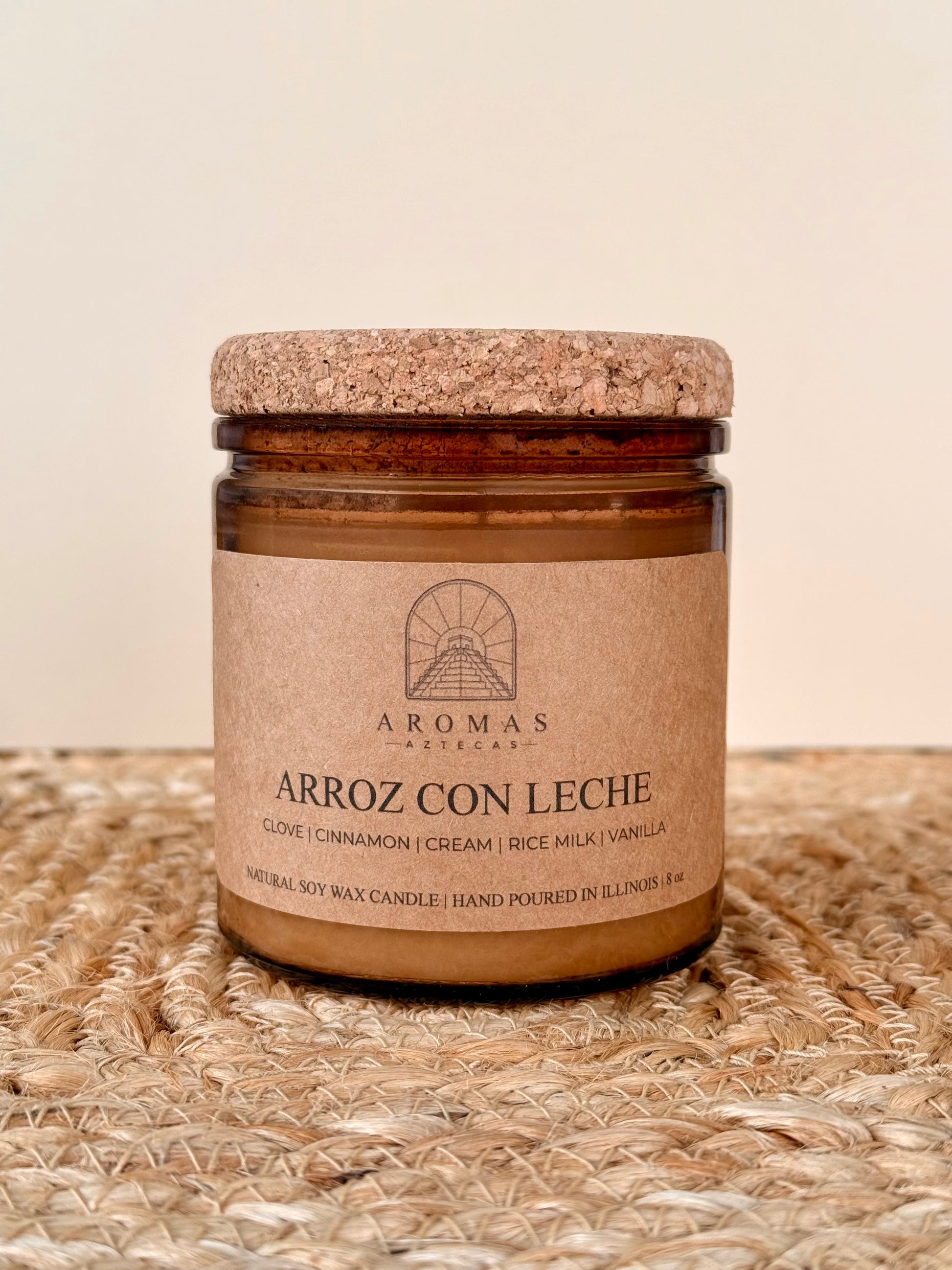 Aromas Aztecas Arroz con Leche soy candle in an amber glass vessel with a cork lid on a woven textured surface.