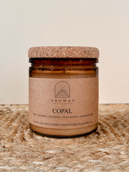 Aromas Aztecas Copal soy candle in an amber glass vessel with a cork lid on a woven textured surface.