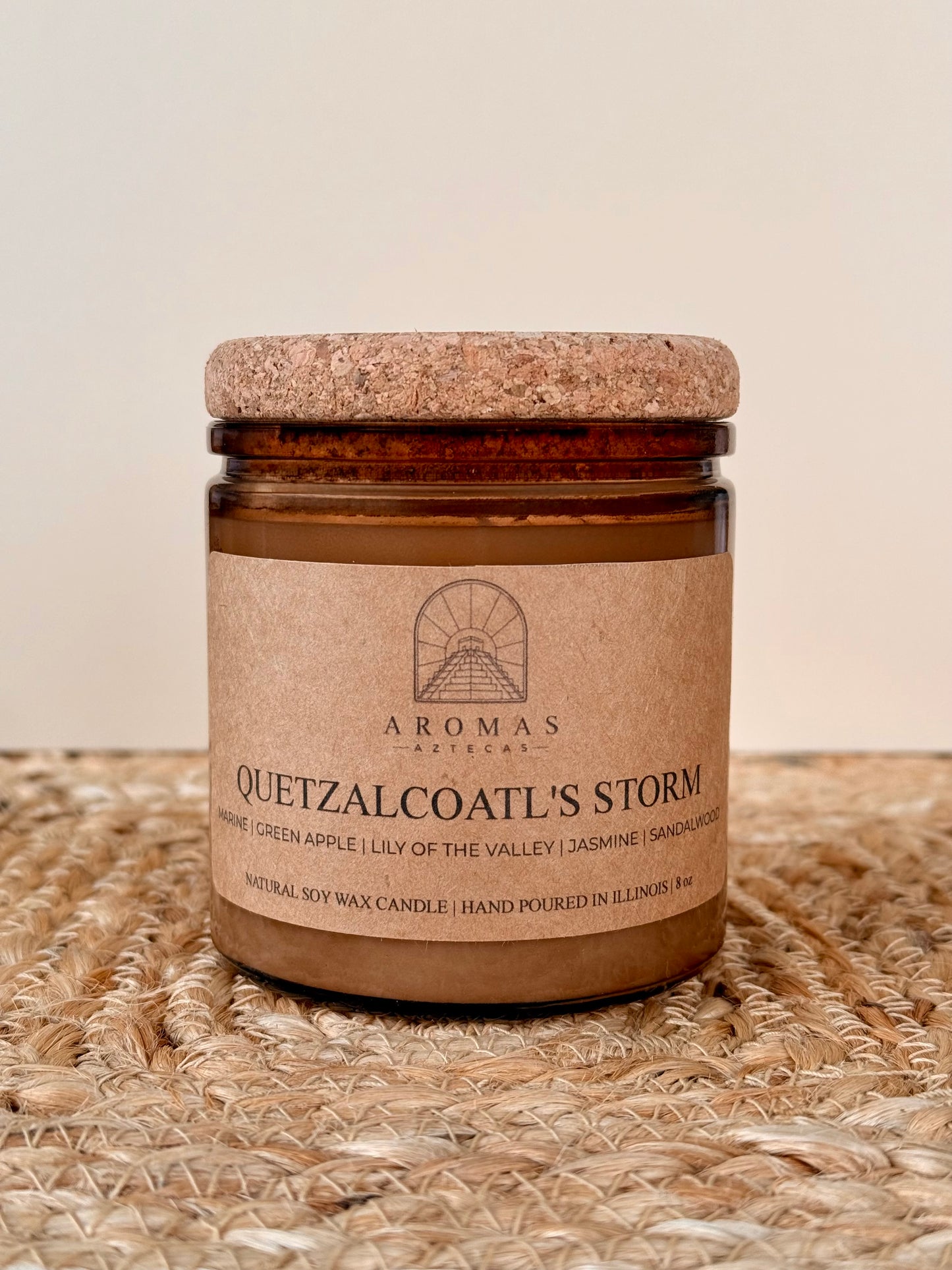 Aromas Aztecas Quetzalcoatl's Storm soy candle in an amber glass vessel with a cork lid on a woven textured surface.