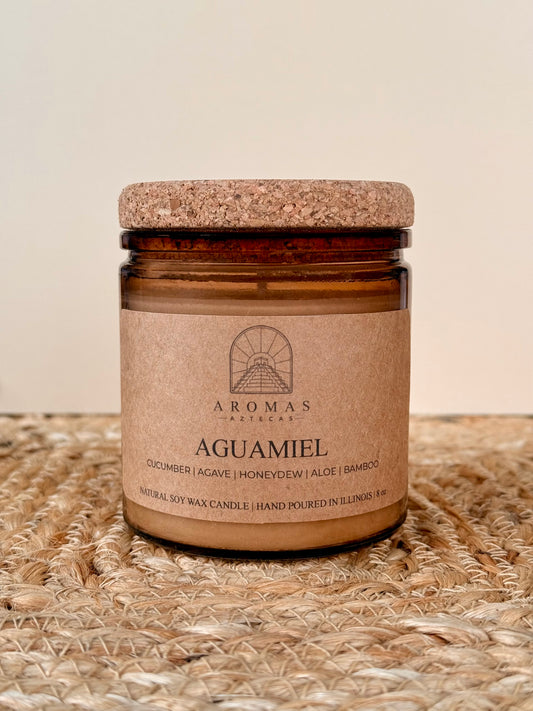 Aromas Aztecas Aguamiel soy candle in an amber glass vessel with a cork lid on a woven textured surface.