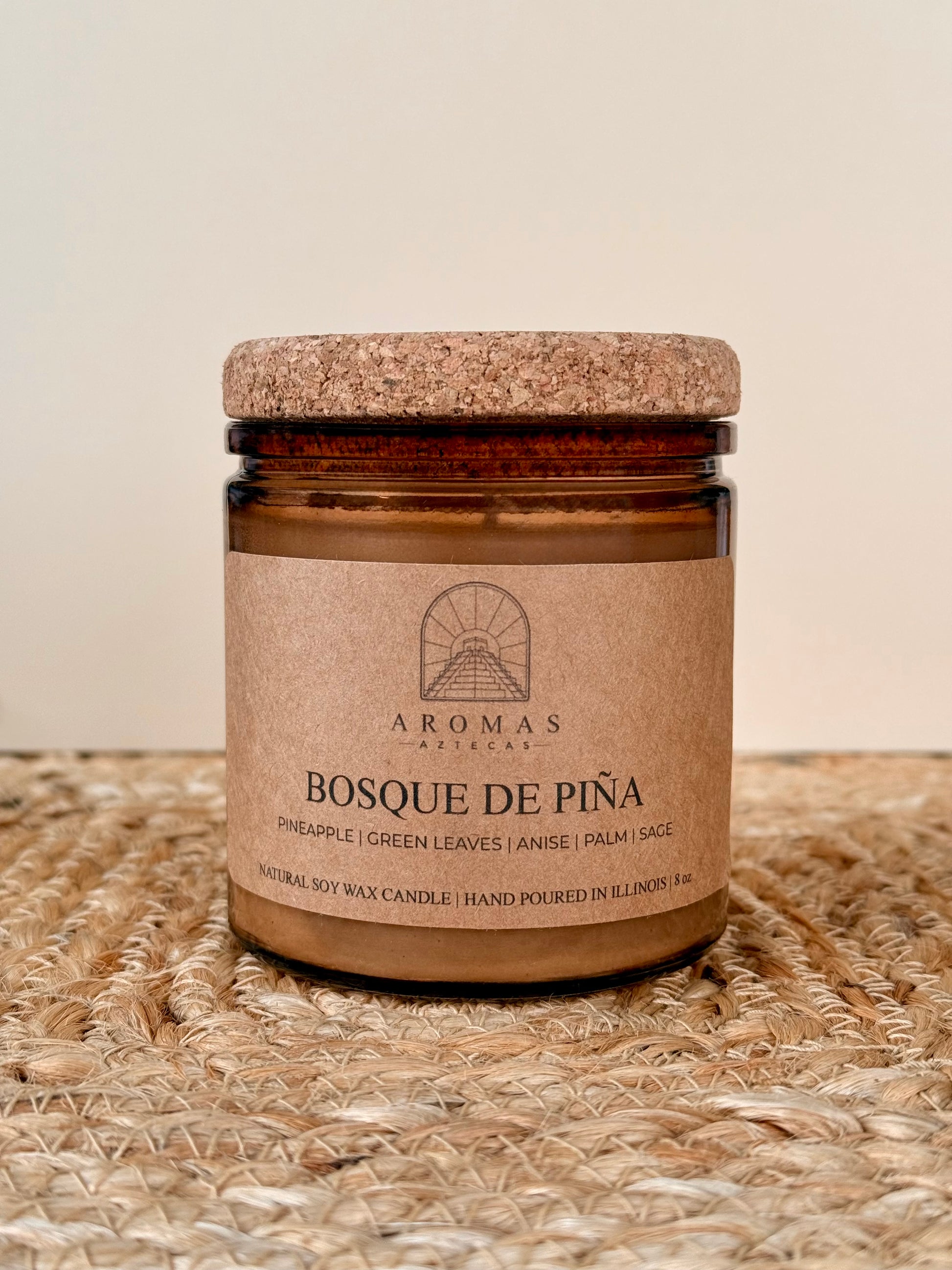 Aromas Aztecas Bosque de Piña soy candle in an amber glass vessel with a cork lid on a woven textured surface.