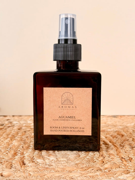 Aromas Aztecas Aguamiel room and linen spray in an amber glass bottle on a woven textured surface.