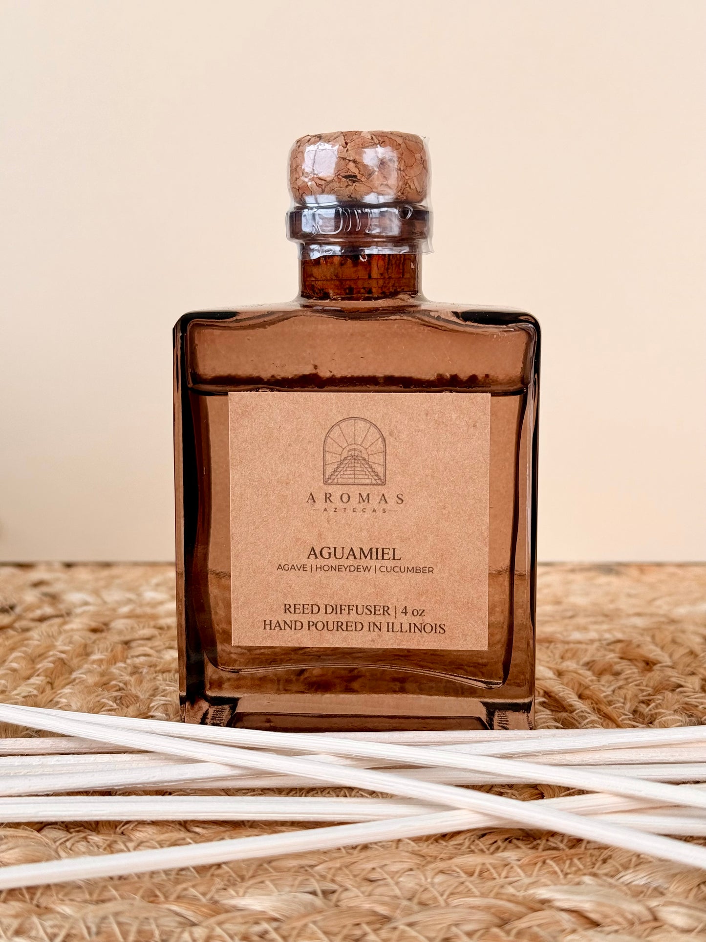 Aromas Aztecas Aguamiel reed diffuser in an amber glass bottle with natural reeds on a woven textured surface.