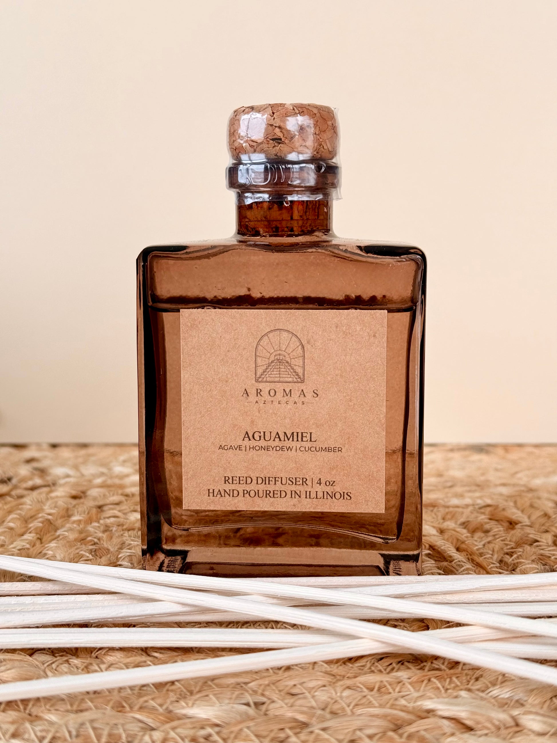Aromas Aztecas Aguamiel reed diffuser in an amber glass bottle with natural reeds on a woven textured surface.
