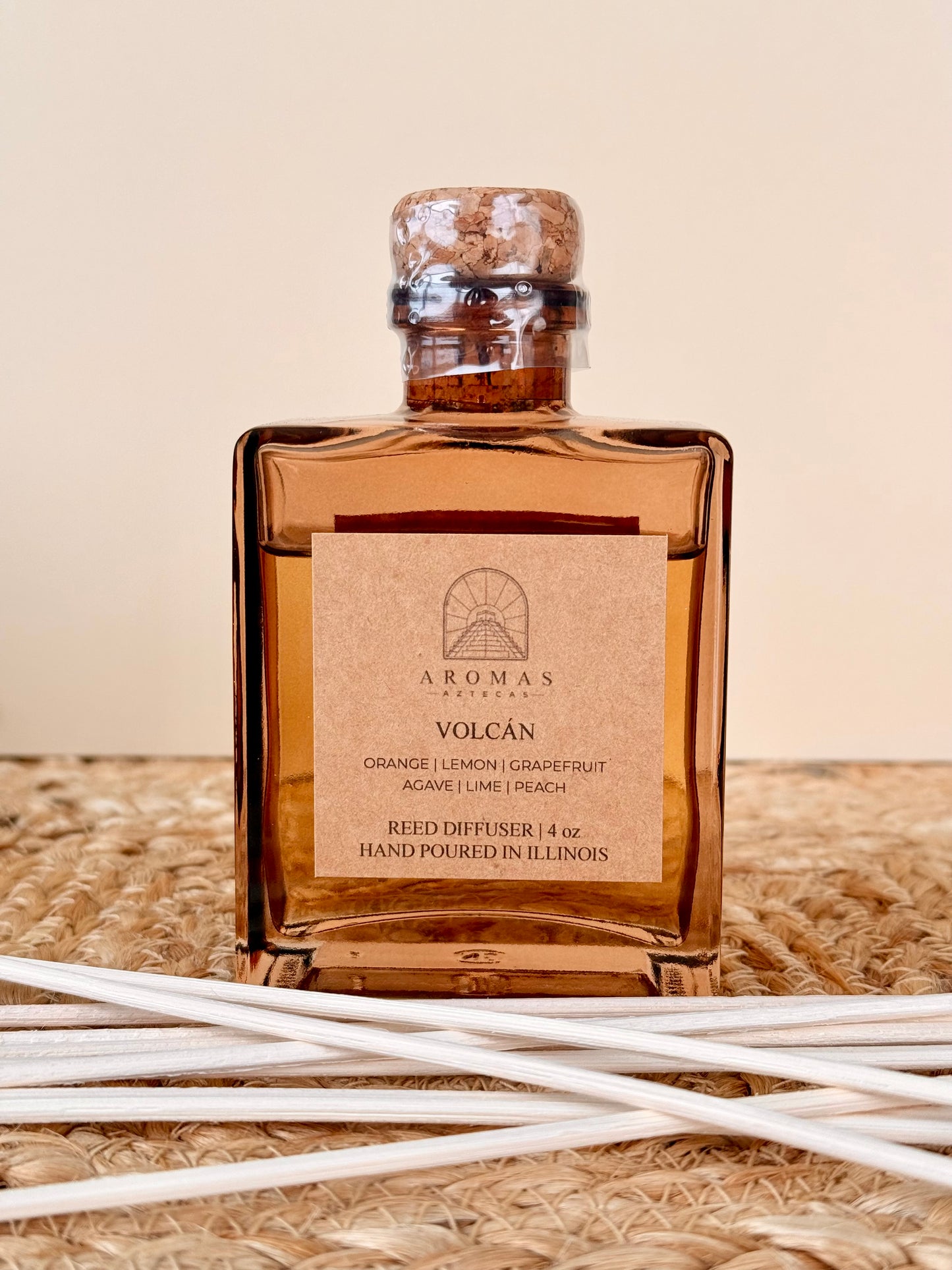Aromas Aztecas Volcán reed diffuser in an amber glass bottle with natural reeds on a woven textured surface.