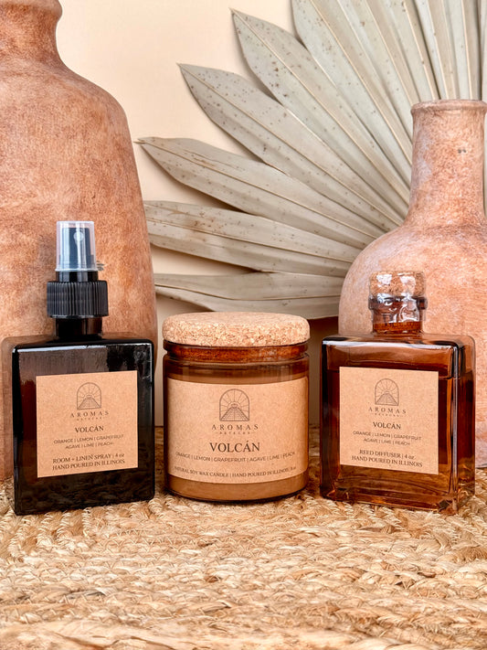 Aromas Aztecas Volcán room and linen spray, soy candle, and reed diffuser in amber glass vessels on a woven textured surface with terracotta pottery and dried palm leaf décor.