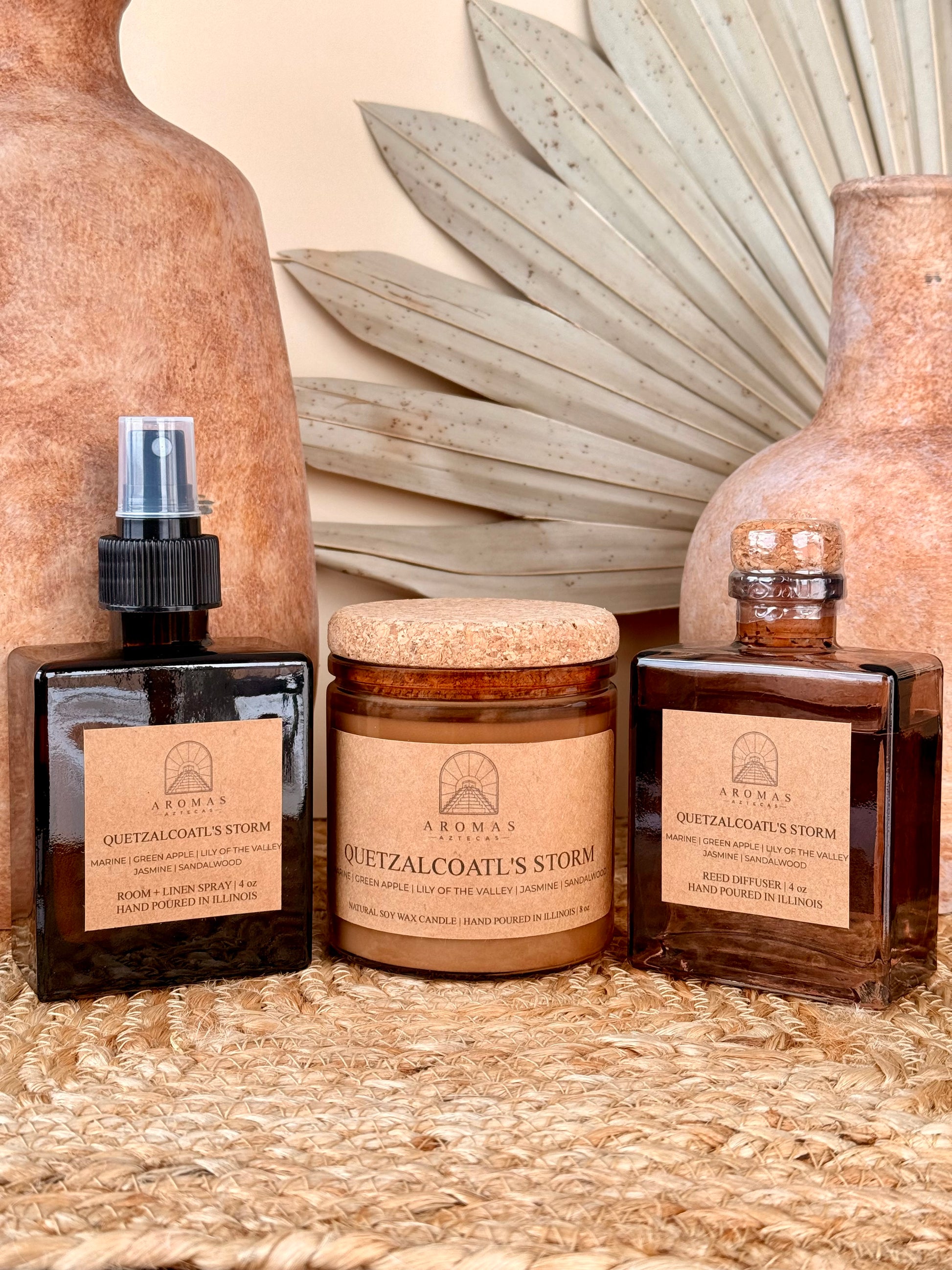 Aromas Aztecas Quetzalcoatl's Storm room and linen spray, soy candle, and reed diffuser in amber glass vessels on a woven textured surface with terracotta pottery and dried palm leaf décor.