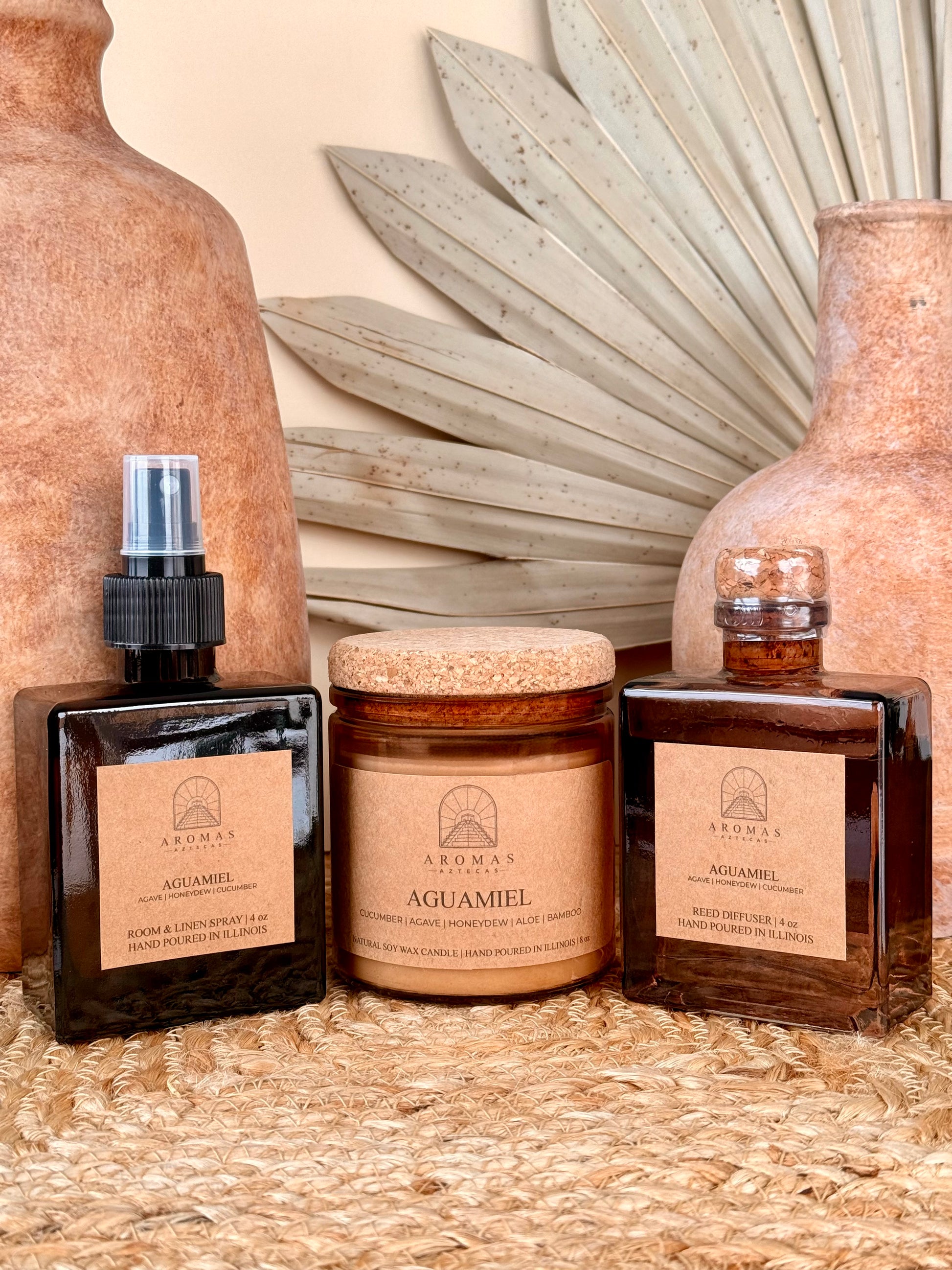 Aromas Aztecas Aguamiel room and linen spray, soy candle, and reed diffuser in amber glass vessels on a woven textured surface with terracotta pottery and dried palm leaf décor.