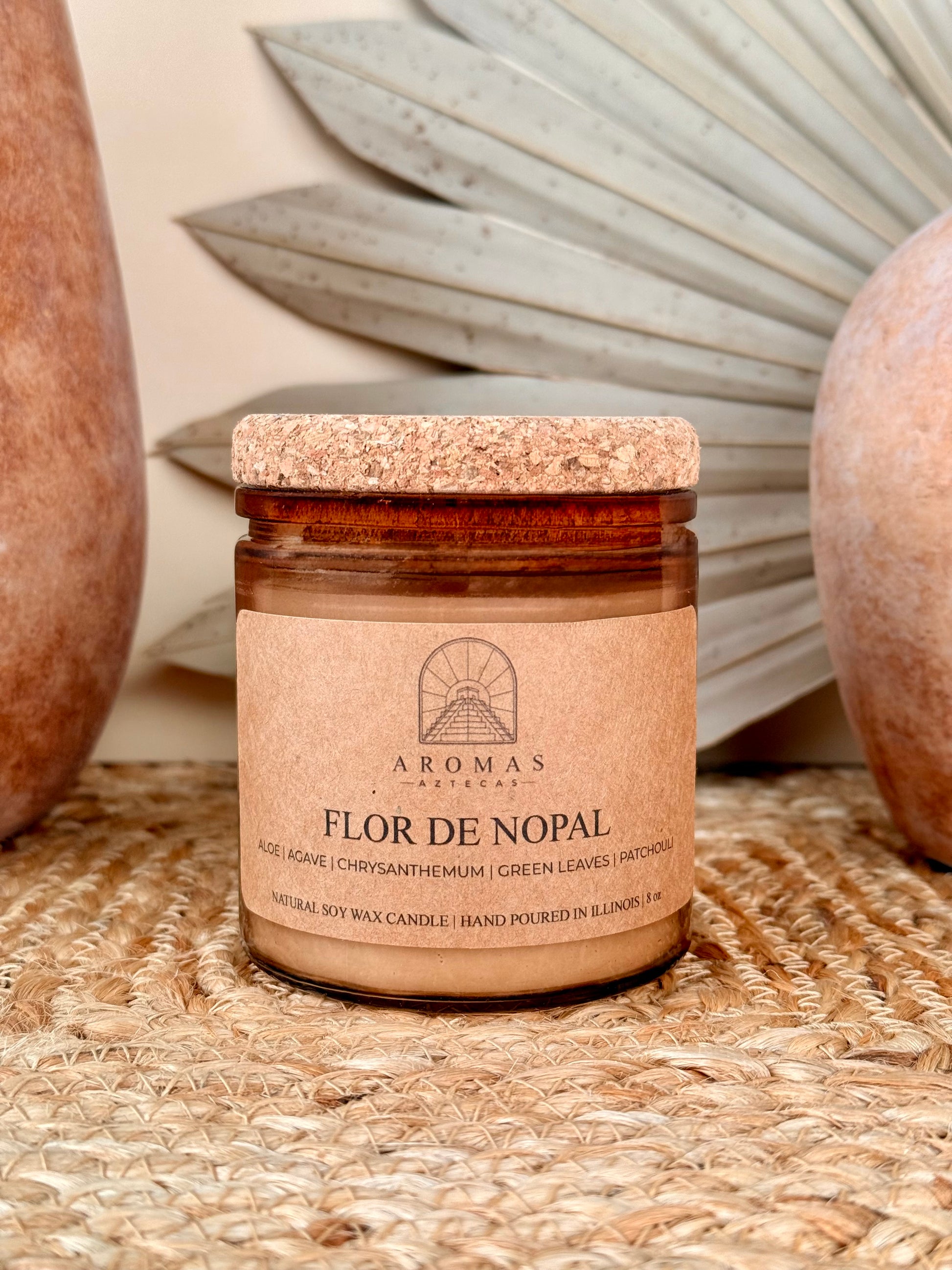 Aromas Aztecas Flor de Nopal soy candle in an amber glass vessel with a cork lid on a woven textured surface with terracotta pottery and dried palm leaf décor.