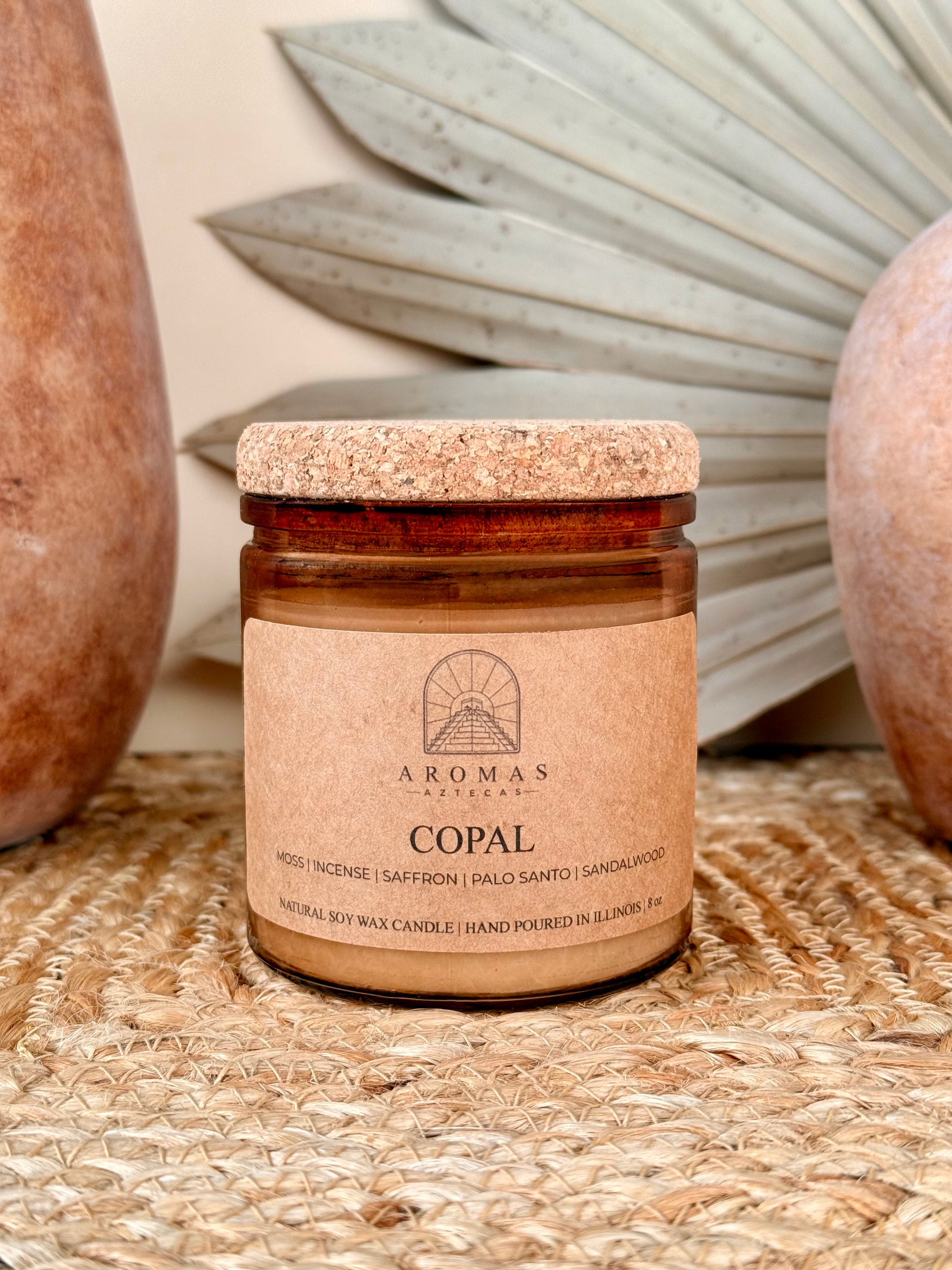 Aromas Aztecas Copal soy candle in an amber glass vessel with a cork lid on a woven textured surface with terracotta pottery and dried palm leaf décor.