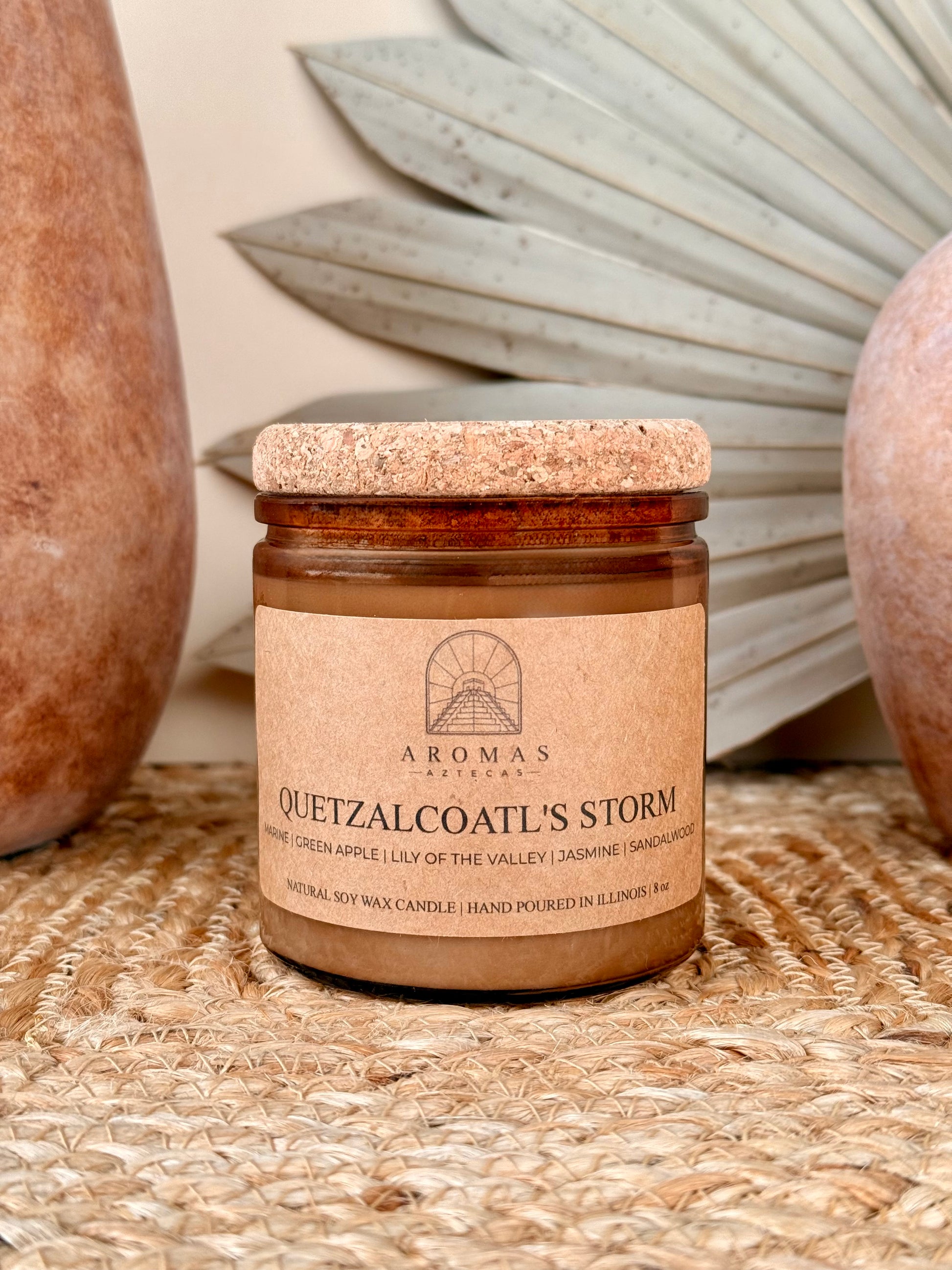 Aromas Aztecas Quetzalcoatl's Storm soy candle in an amber glass vessel with a cork lid on a woven textured surface with terracotta pottery and dried palm leaf décor.