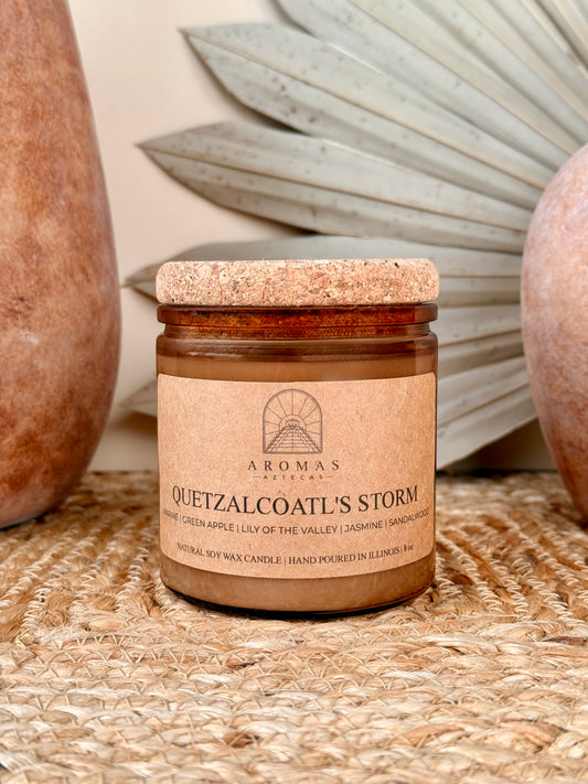 Aromas Aztecas Quetzalcoatl's Storm soy candle in an amber glass vessel with a cork lid on a woven textured surface with terracotta pottery and dried palm leaf décor.