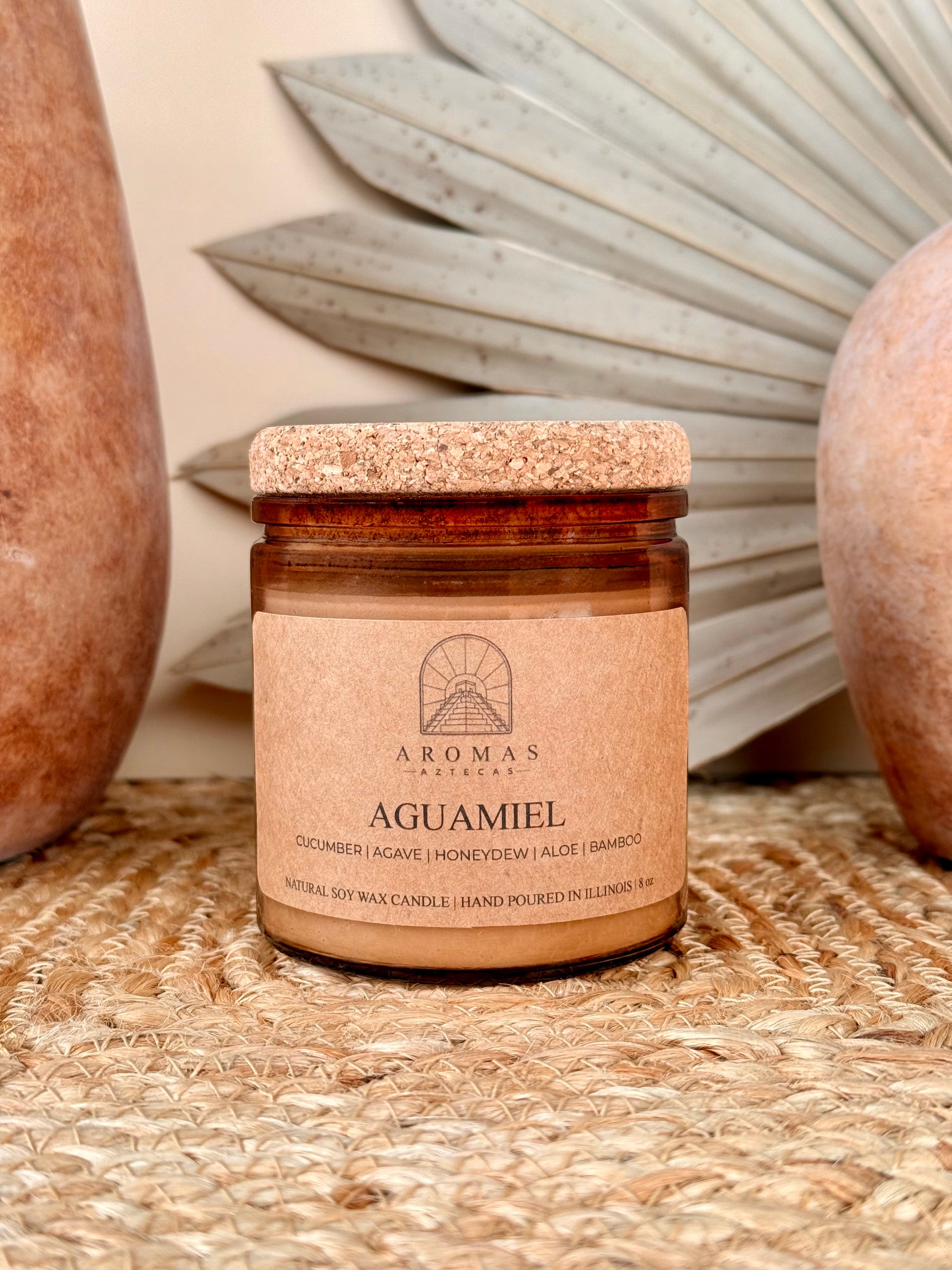 Aromas Aztecas Aguamiel soy candle in an amber glass vessel with a cork lid on a woven textured surface with terracotta pottery and dried palm leaf décor.