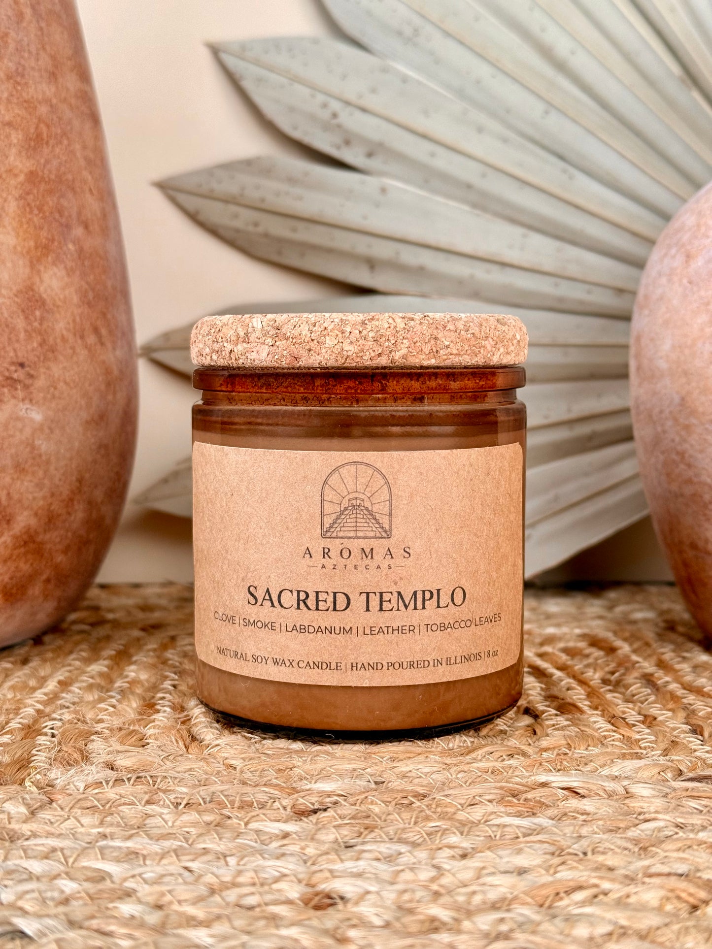 Aromas Aztecas Sacred Templo soy candle in an amber glass vessel with a cork lid on a woven textured surface with terracotta pottery and dried palm leaf décor.