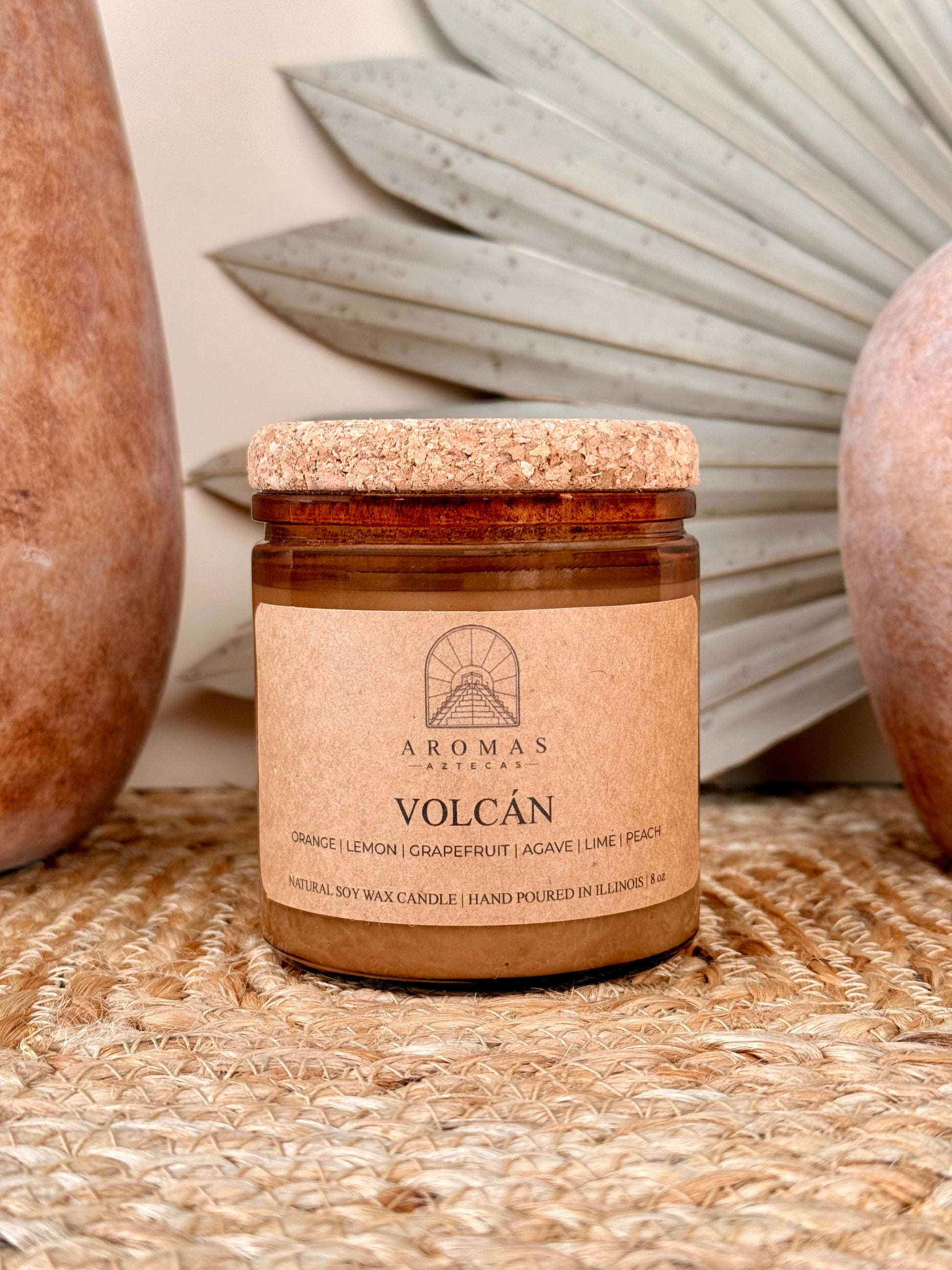 Aromas Aztecas Volcán soy candle in an amber glass vessel with a cork lid on a woven textured surface with terracotta pottery and dried palm leaf décor.