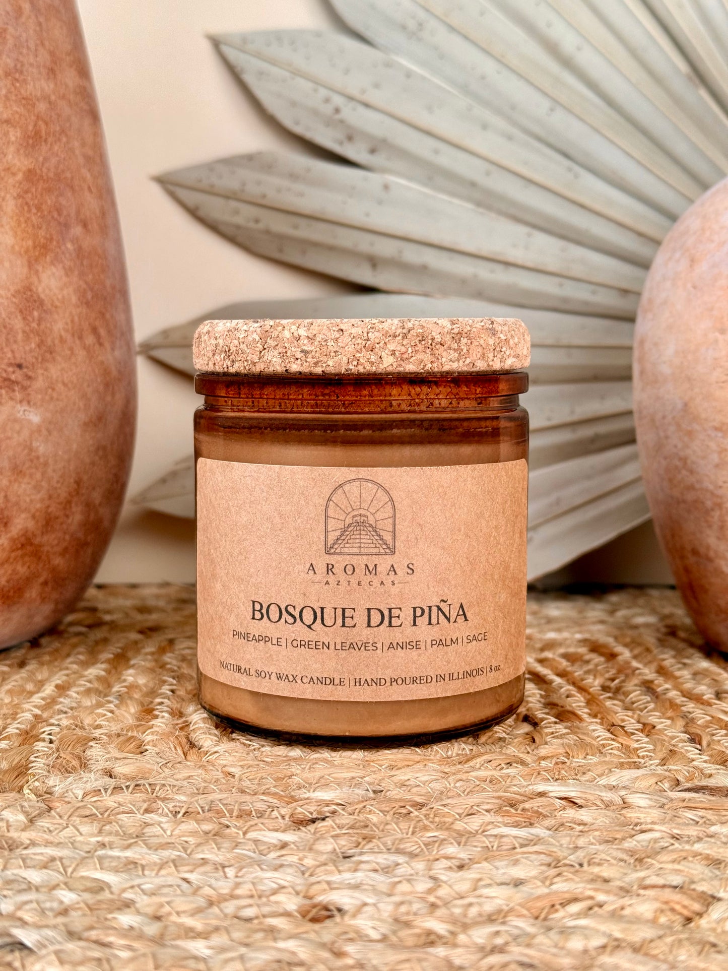 Aromas Aztecas Bosque de Piña soy candle in an amber glass vessel with a cork lid on a woven textured surface with terracotta pottery and dried palm leaf décor.