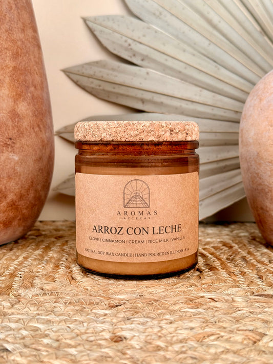 Aromas Aztecas Arroz con Leche soy candle in an amber glass vessel with a cork lid on a woven textured surface with terracotta pottery and dried palm leaf décor.