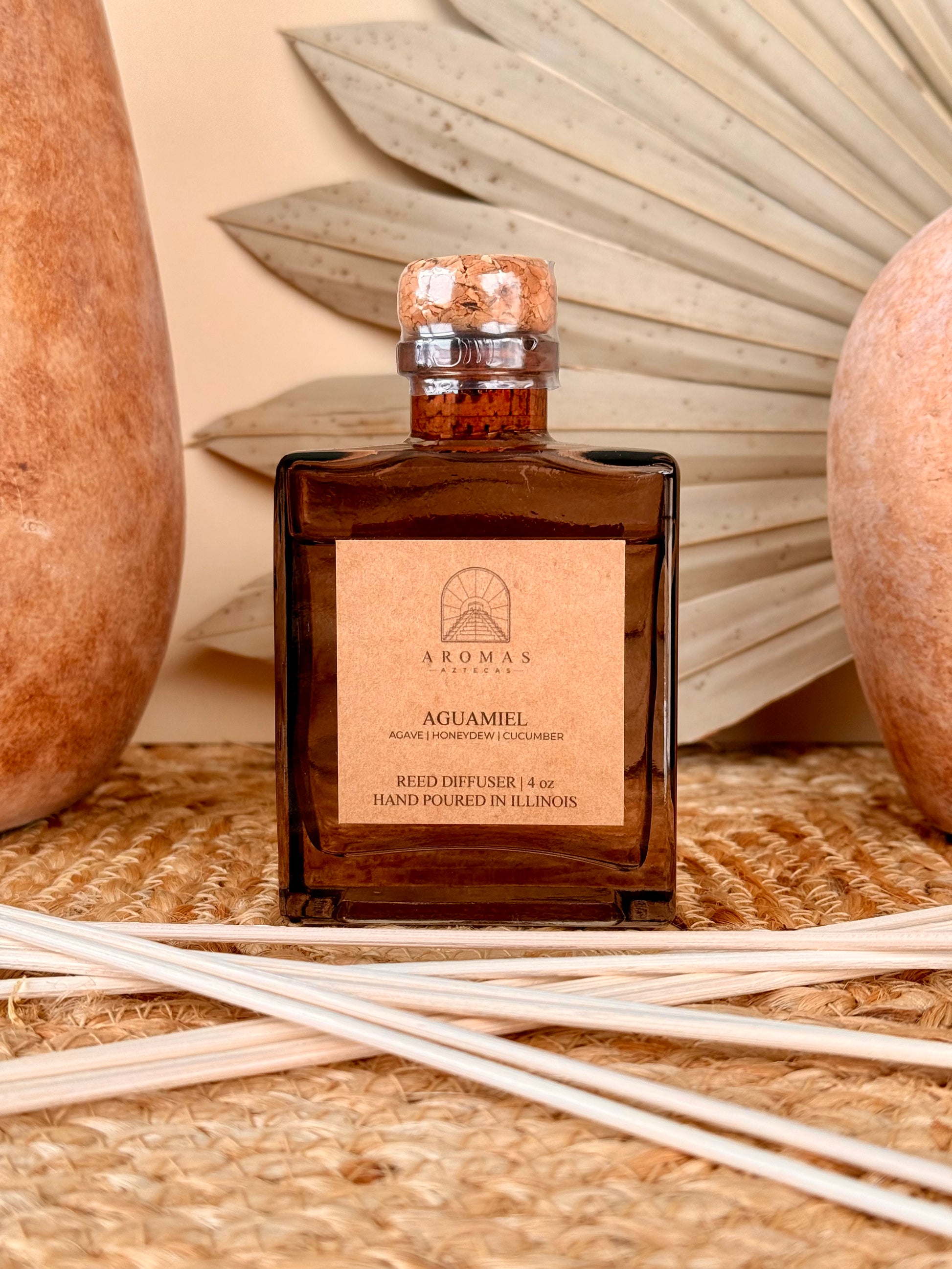 Aromas Aztecas Aguamiel reed diffuser in an amber glass bottle with natural reeds on a woven textured surface with terracotta pottery and dried palm leaf décor.