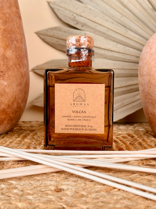 Aromas Aztecas Volcán reed diffuser in an amber glass bottle with natural reeds on a woven textured surface with terracotta pottery and dried palm leaf décor.