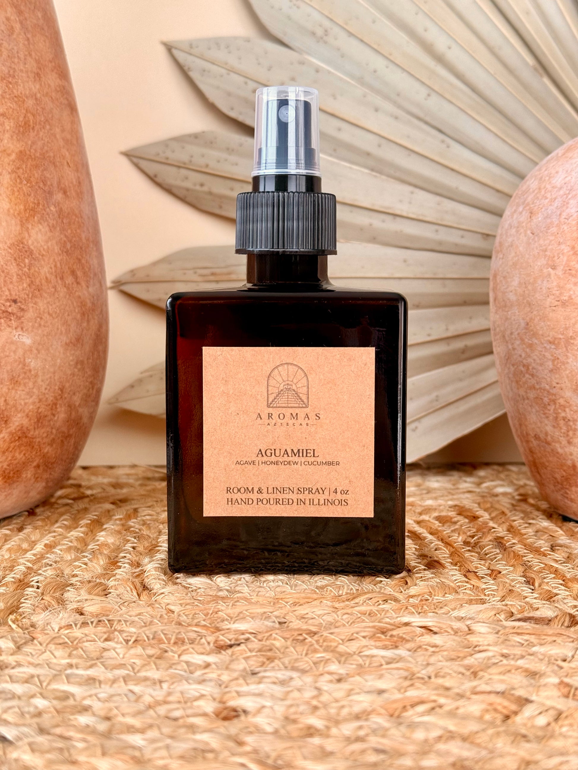 Aromas Aztecas Aguamiel room and linen spray in an amber glass bottle on a woven textured surface with terracotta pottery and dried palm leaf décor.