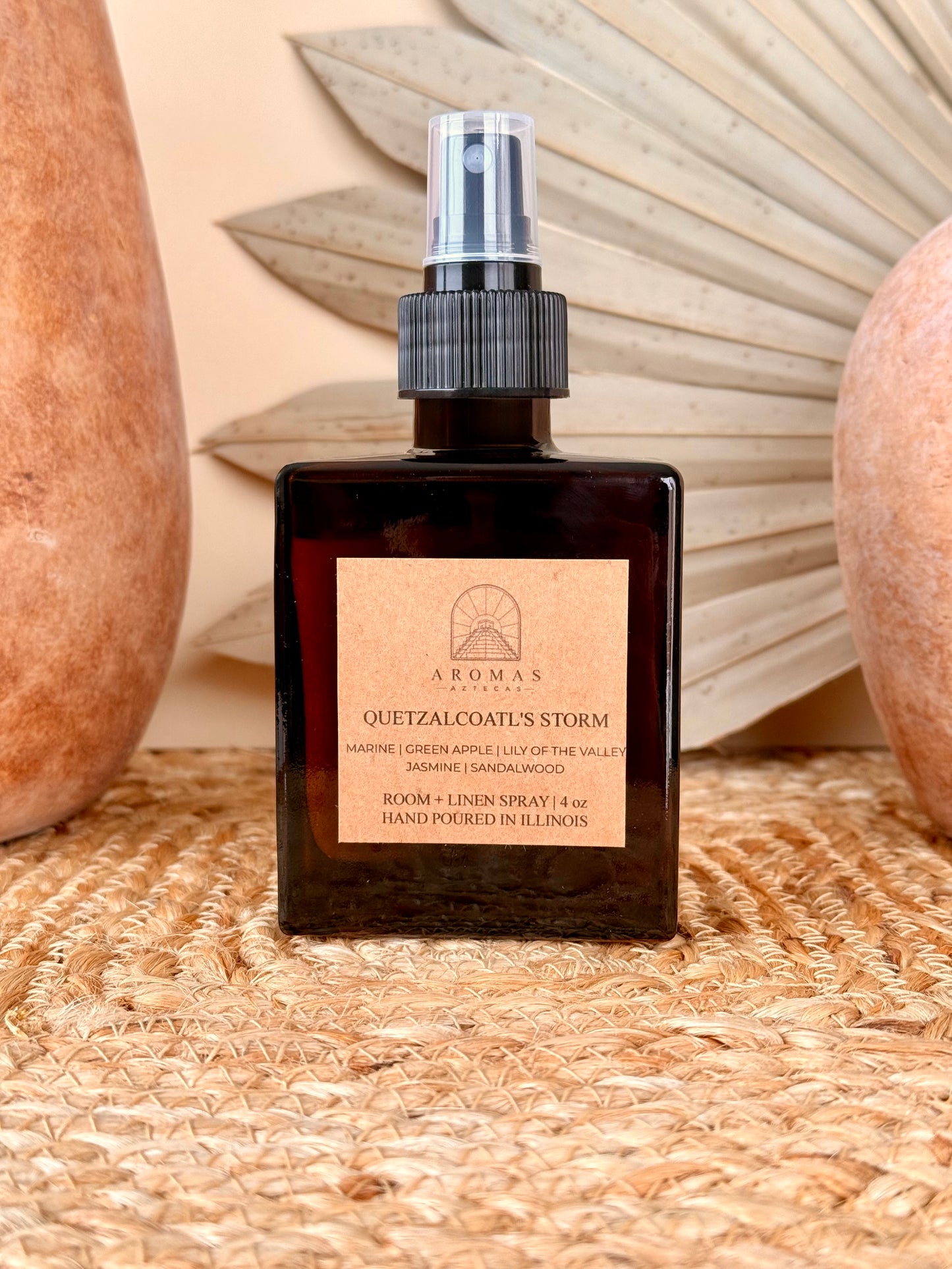 Aromas Aztecas Quetzalcoatl's Storm room and linen spray in an amber glass bottle on a woven textured surface with terracotta pottery and dried palm leaf décor.
