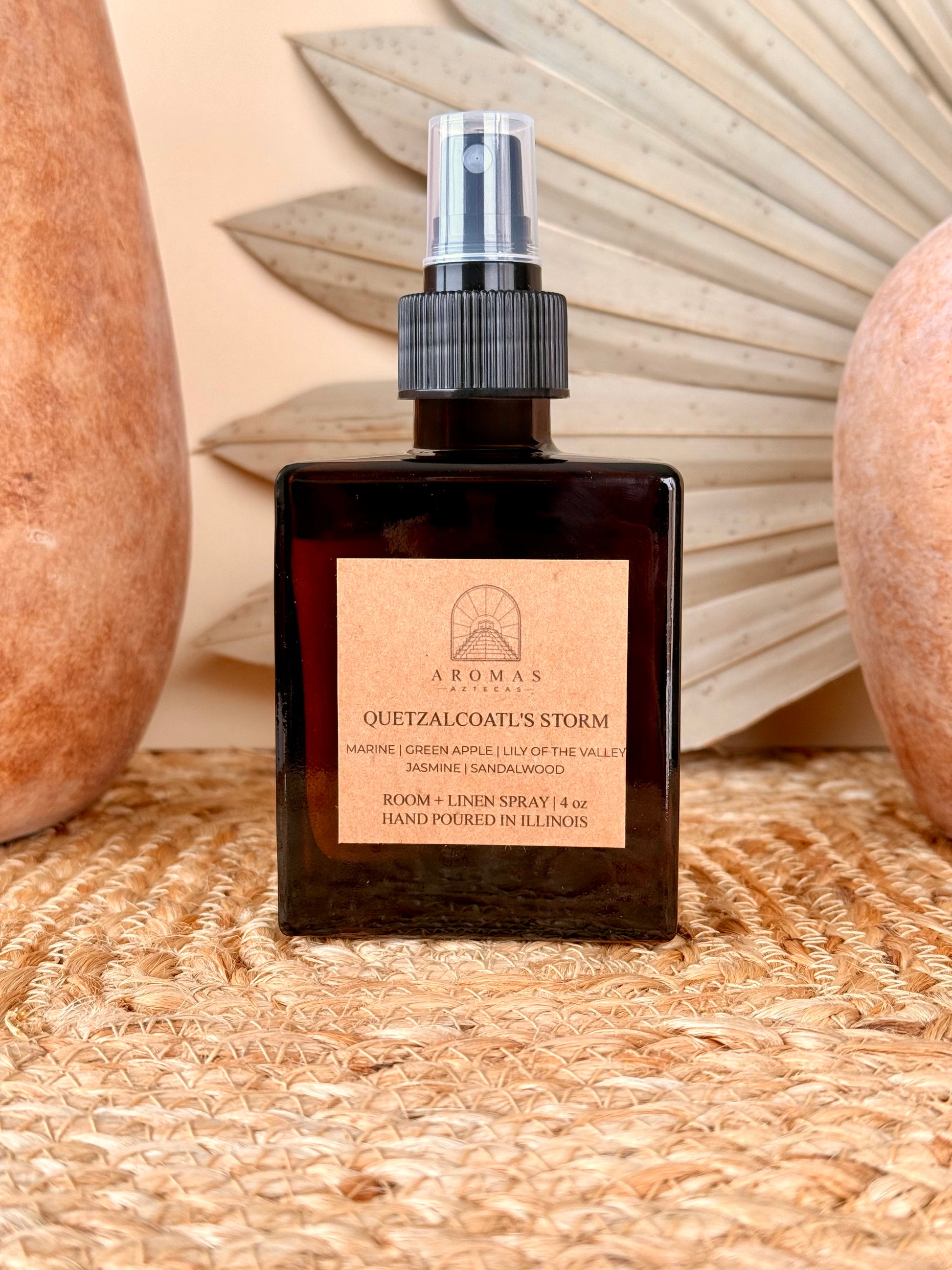 Aromas Aztecas Quetzalcoatl's Storm room and linen spray in an amber glass bottle on a woven textured surface with terracotta pottery and dried palm leaf décor.
