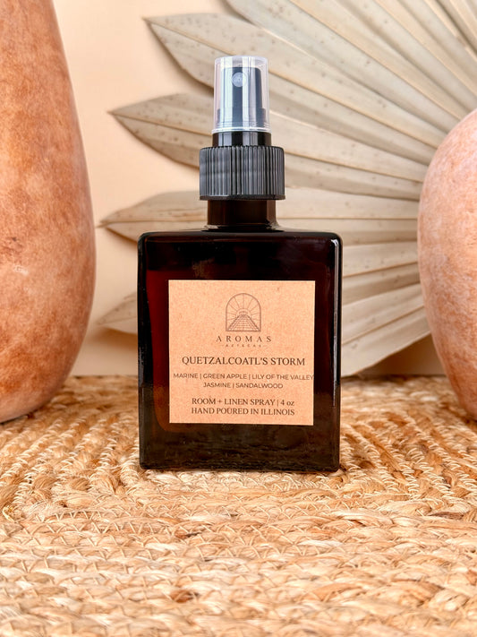 Aromas Aztecas Quetzalcoatl's Storm room and linen spray in an amber glass bottle on a woven textured surface with terracotta pottery and dried palm leaf décor.