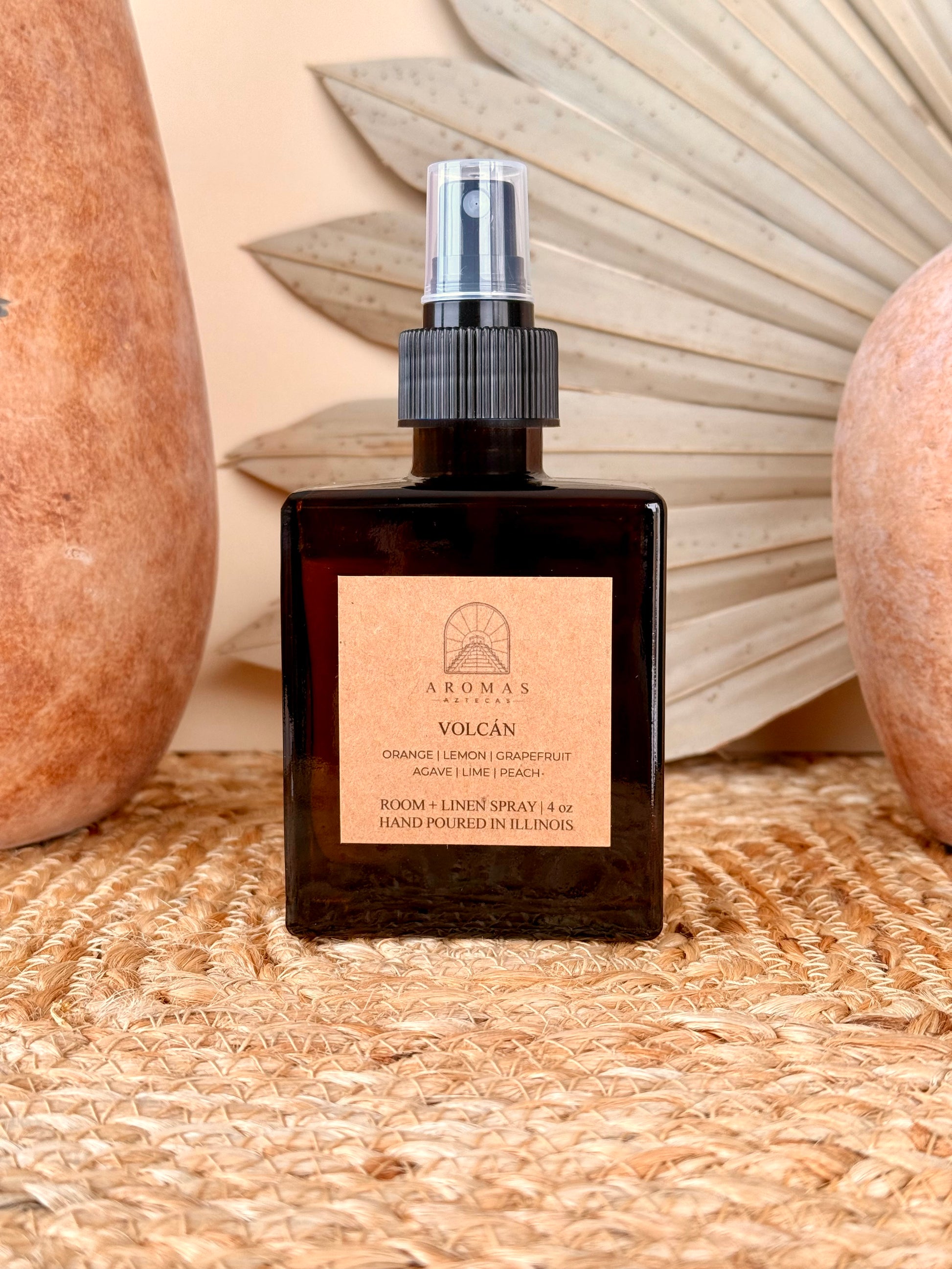 Aromas Aztecas Volcán room and linen spray in an amber glass bottle on a woven textured surface with terracotta pottery and dried palm leaf décor.