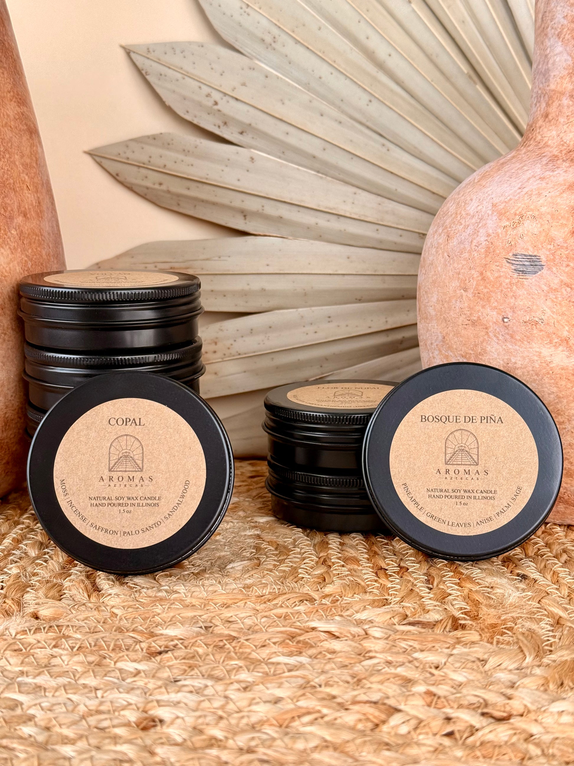 Aromas Aztecas discovery soy candle set with multiple fragrance samples featuring Copal and Bosque de Piña in black aluminum tins on a woven textured surface with terracotta pottery and dried palm leaf décor.