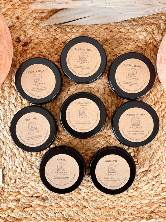 Aromas Aztecas discovery soy candle set with multiple fragrance samples in black aluminum tins on a woven textured surface with terracotta pottery and dried palm leaf décor.