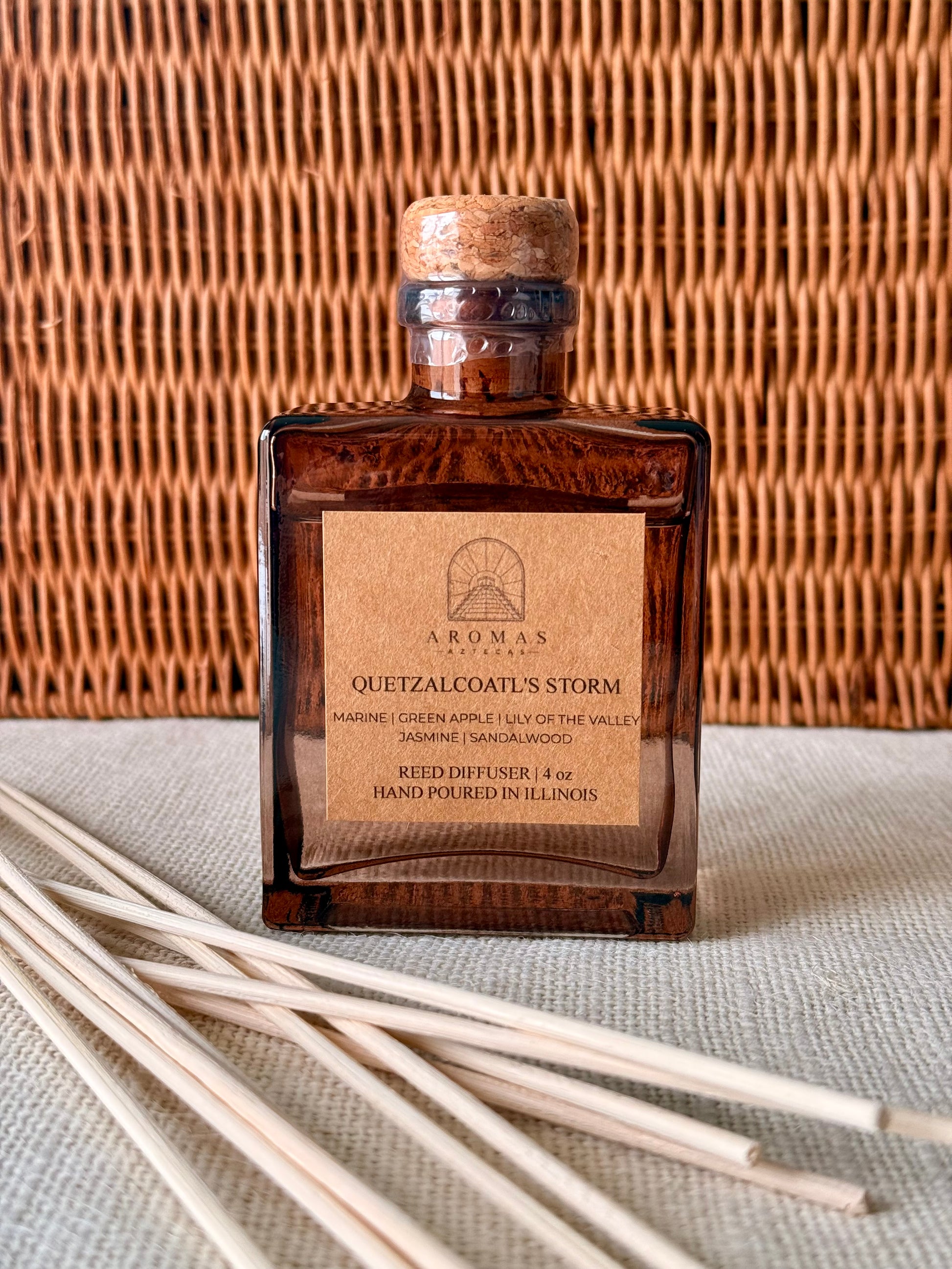 Aromas Aztecas Quetzalcoatl's Storm reed diffuser in an amber glass bottle with natural reeds against a woven textured backdrop.