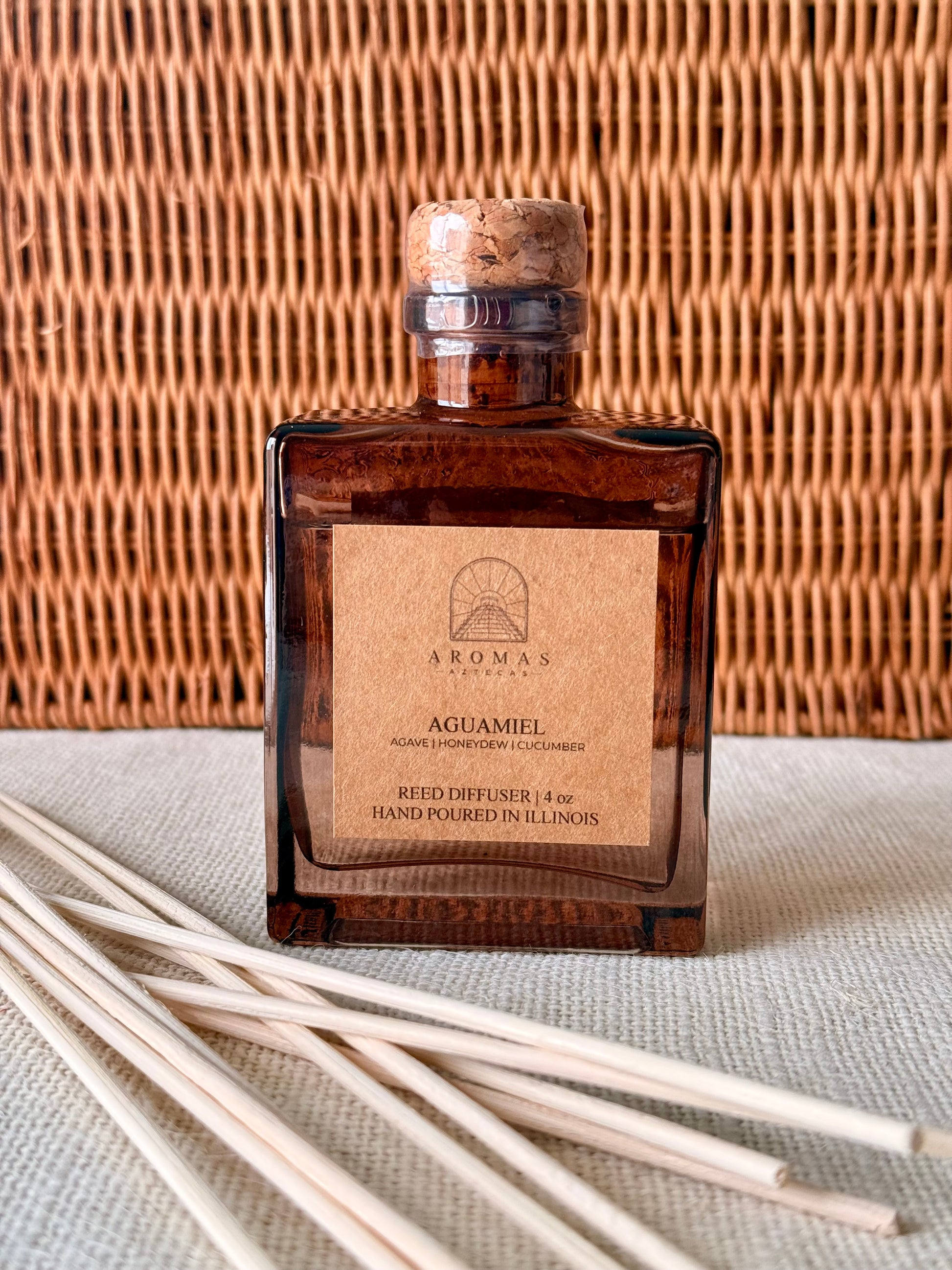 Aromas Aztecas Aguamiel reed diffuser in an amber glass bottle with natural reeds against a woven textured surface.