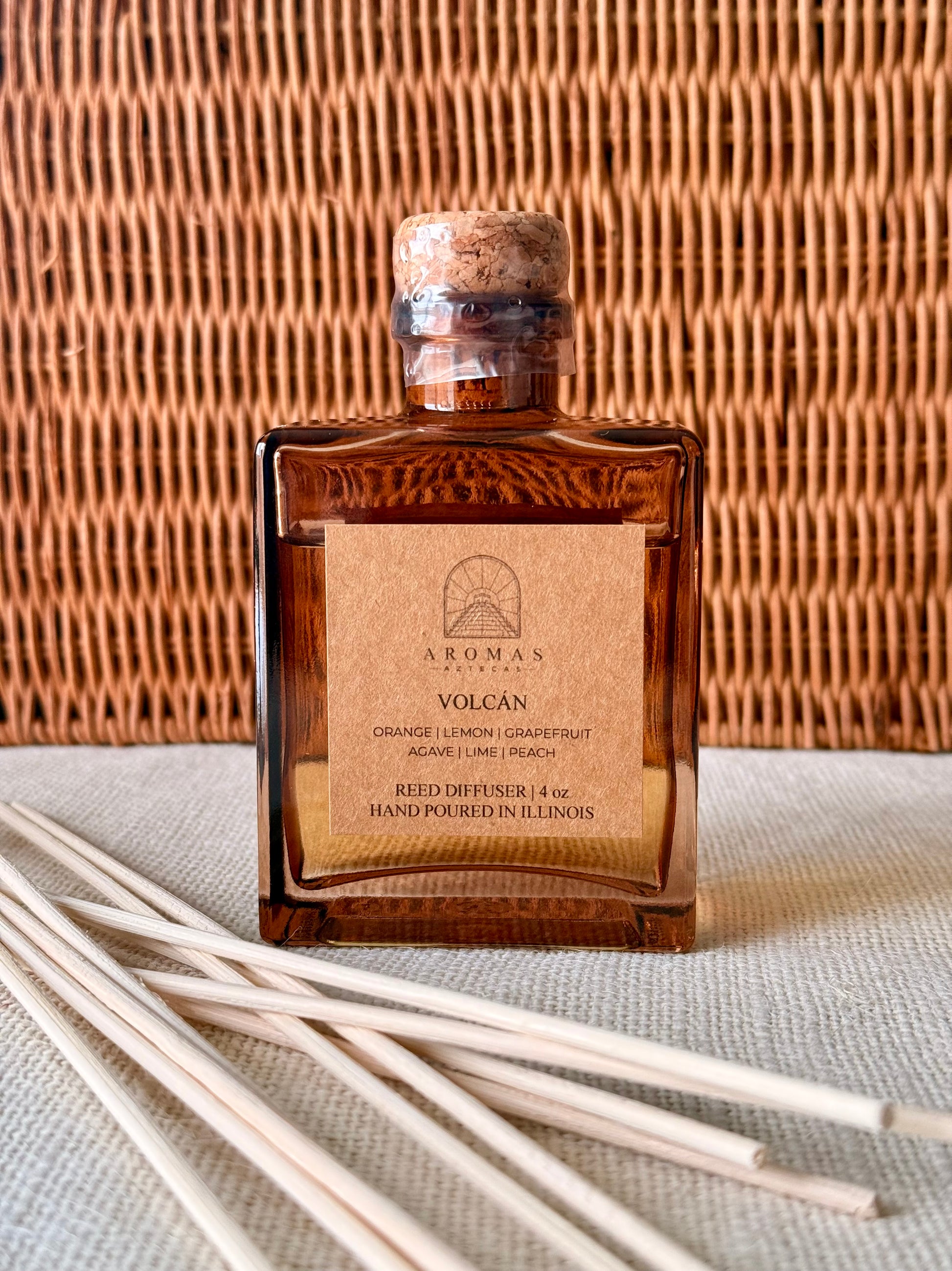 Aromas Aztecas Volcán reed diffuser in an amber glass bottle with natural reeds and woven textured backdrop.