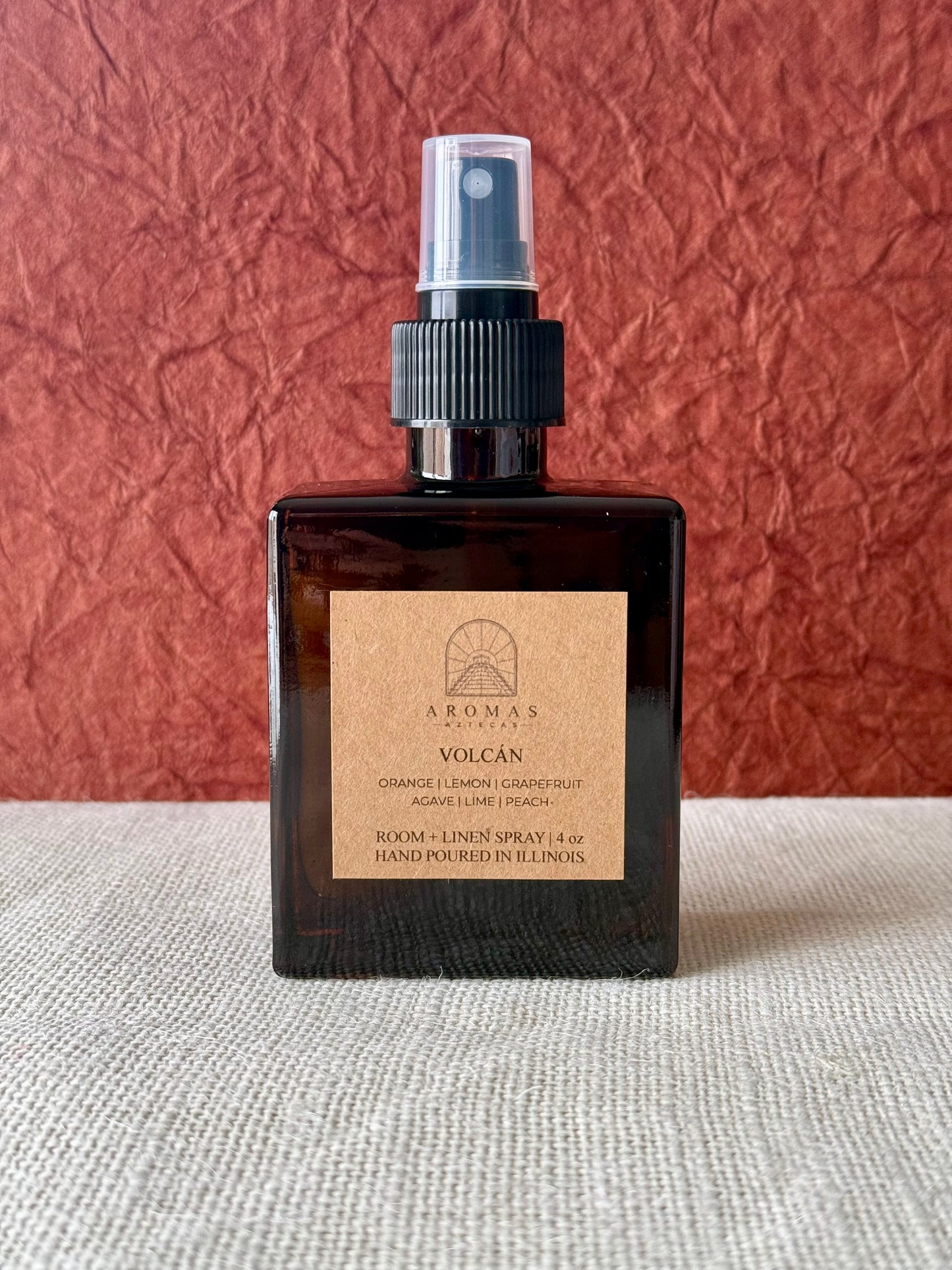 Aromas Aztecas Volcán room and linen spray in an amber glass bottle on a woven textured surface with a burgundy textured backdrop.