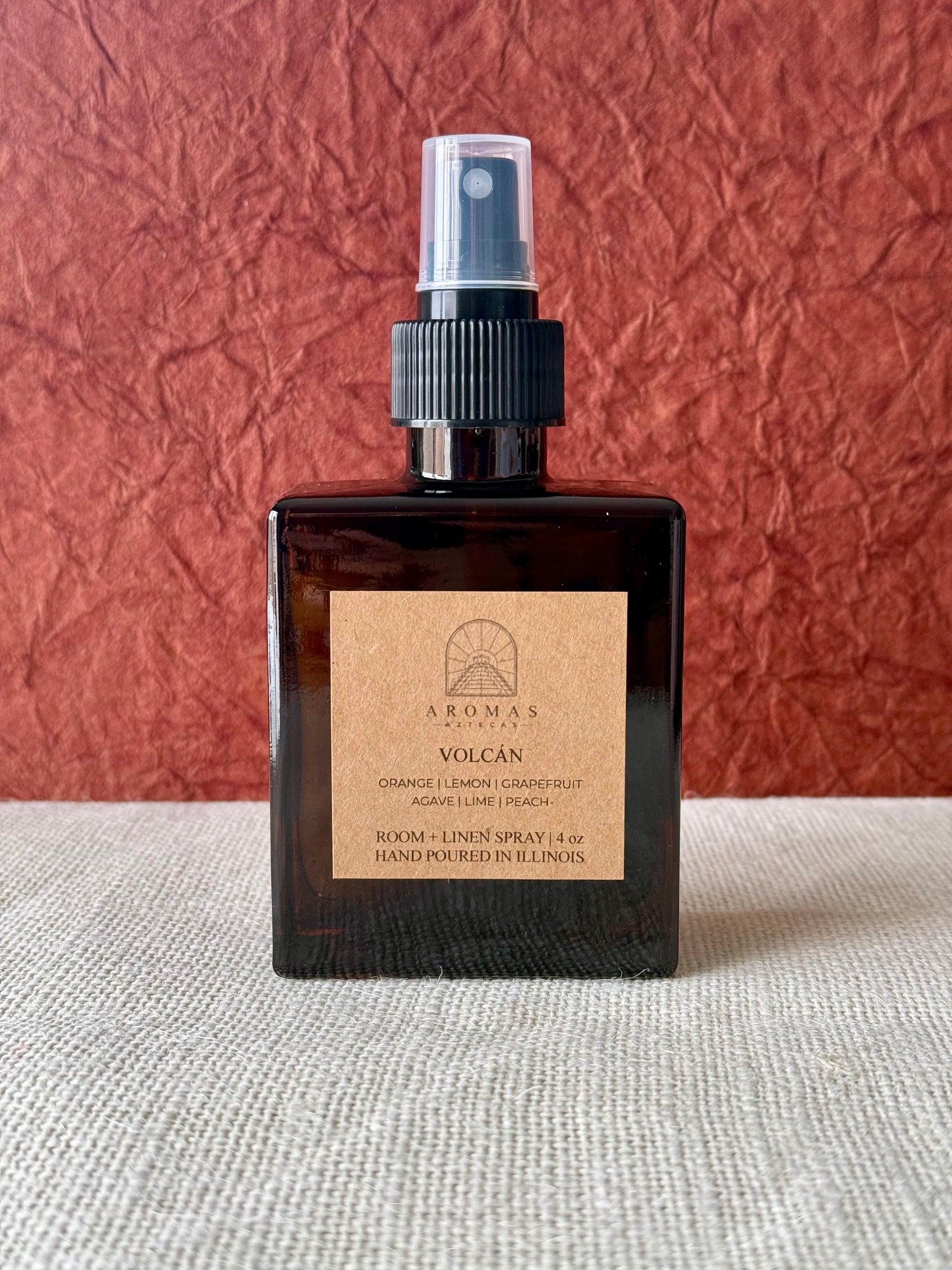 Aromas Aztecas Volcán room and linen spray in an amber glass bottle on a woven textured surface with a burgundy textured backdrop.