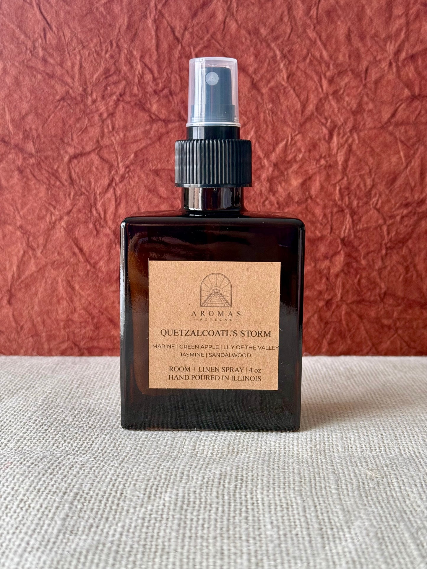 Aromas Aztecas Quetzalcoatl's Storm room and linen spray in an amber glass bottle on a woven textured surface and a burgundy textured backdrop.