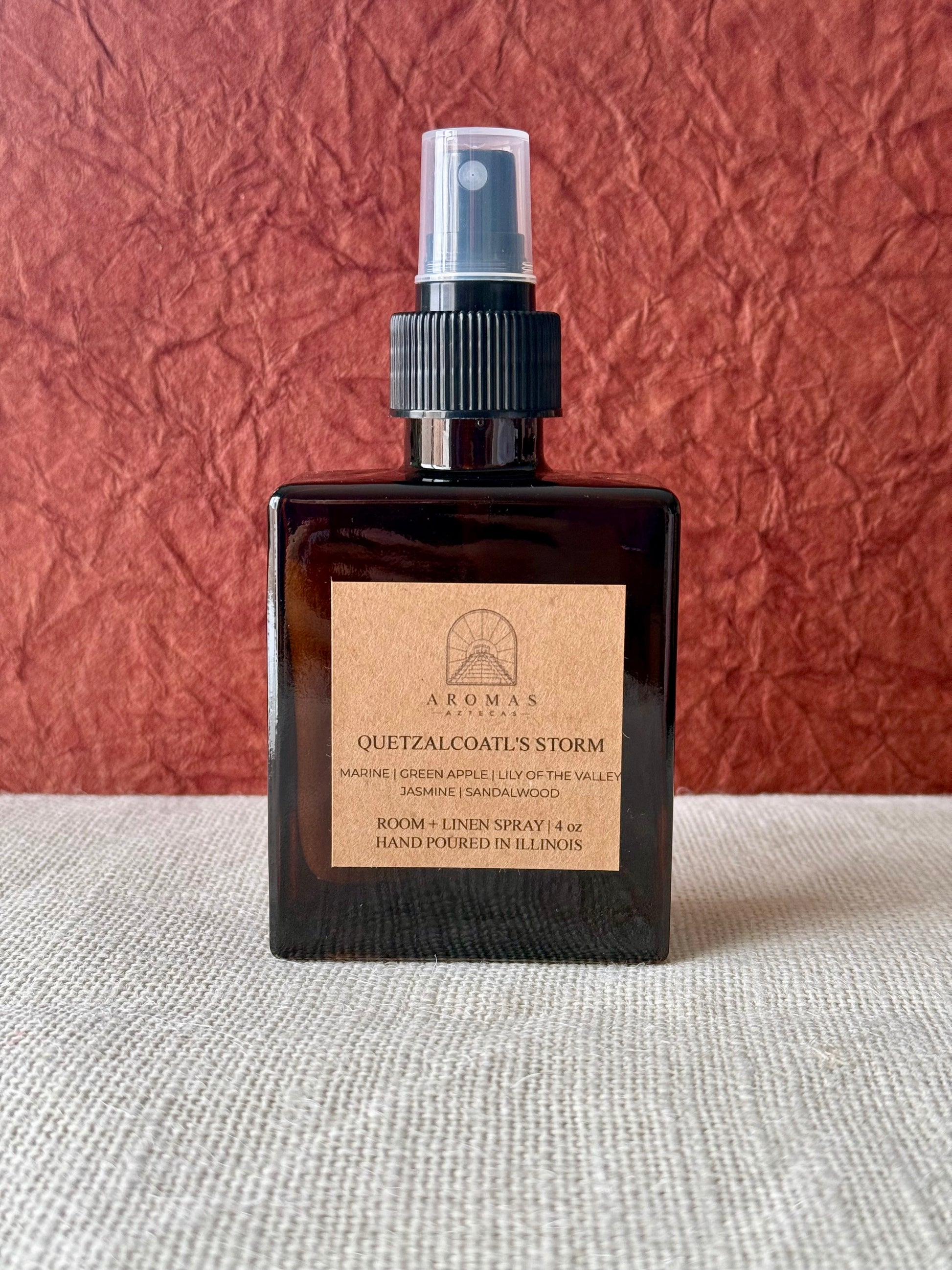 Aromas Aztecas Quetzalcoatl's Storm room and linen spray in an amber glass bottle on a woven textured surface and a burgundy textured backdrop.