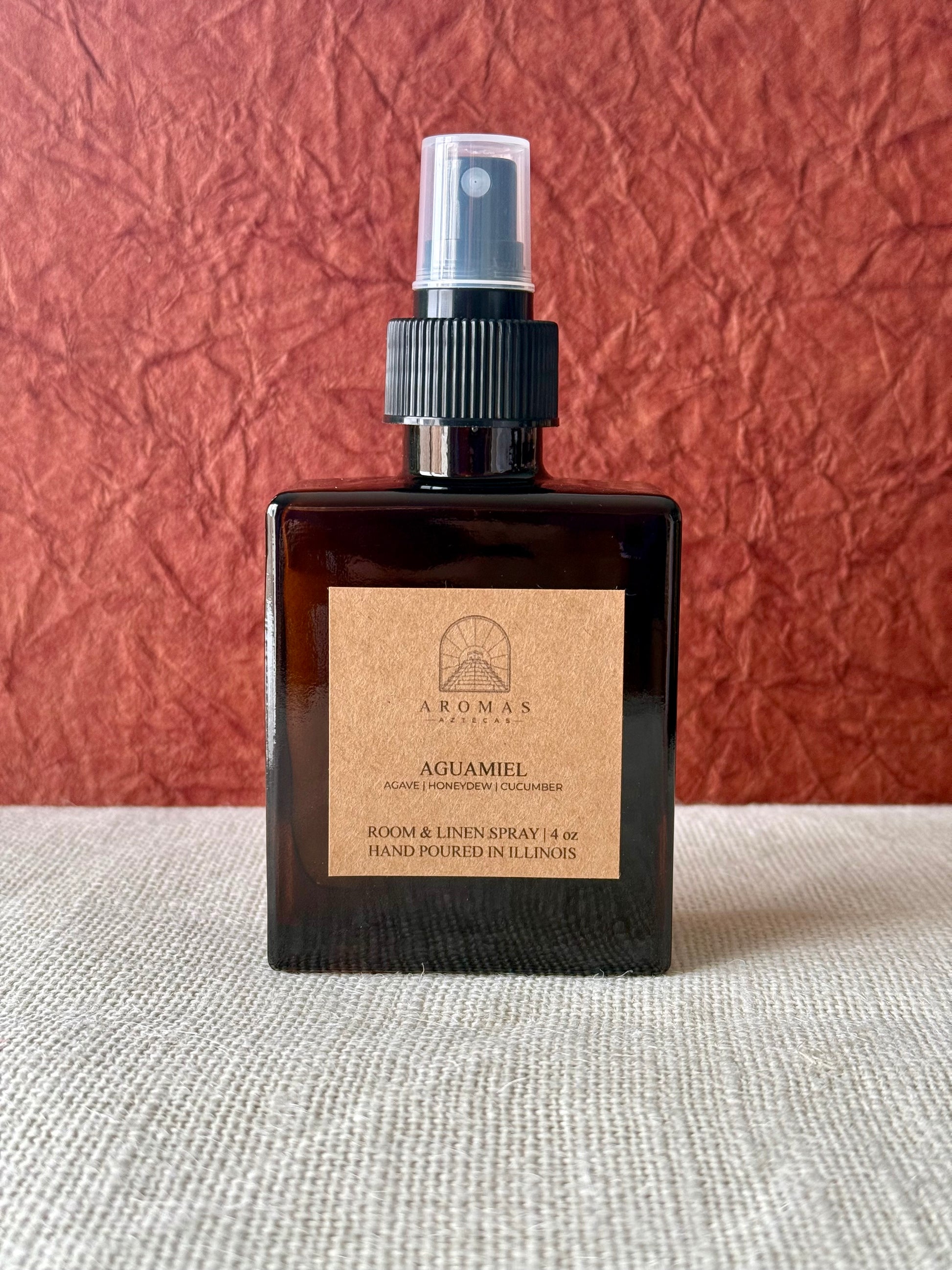 Aromas Aztecas Aguamiel room and linen spray in an amber glass bottle on a woven textured surface and a burgundy textured backdrop.